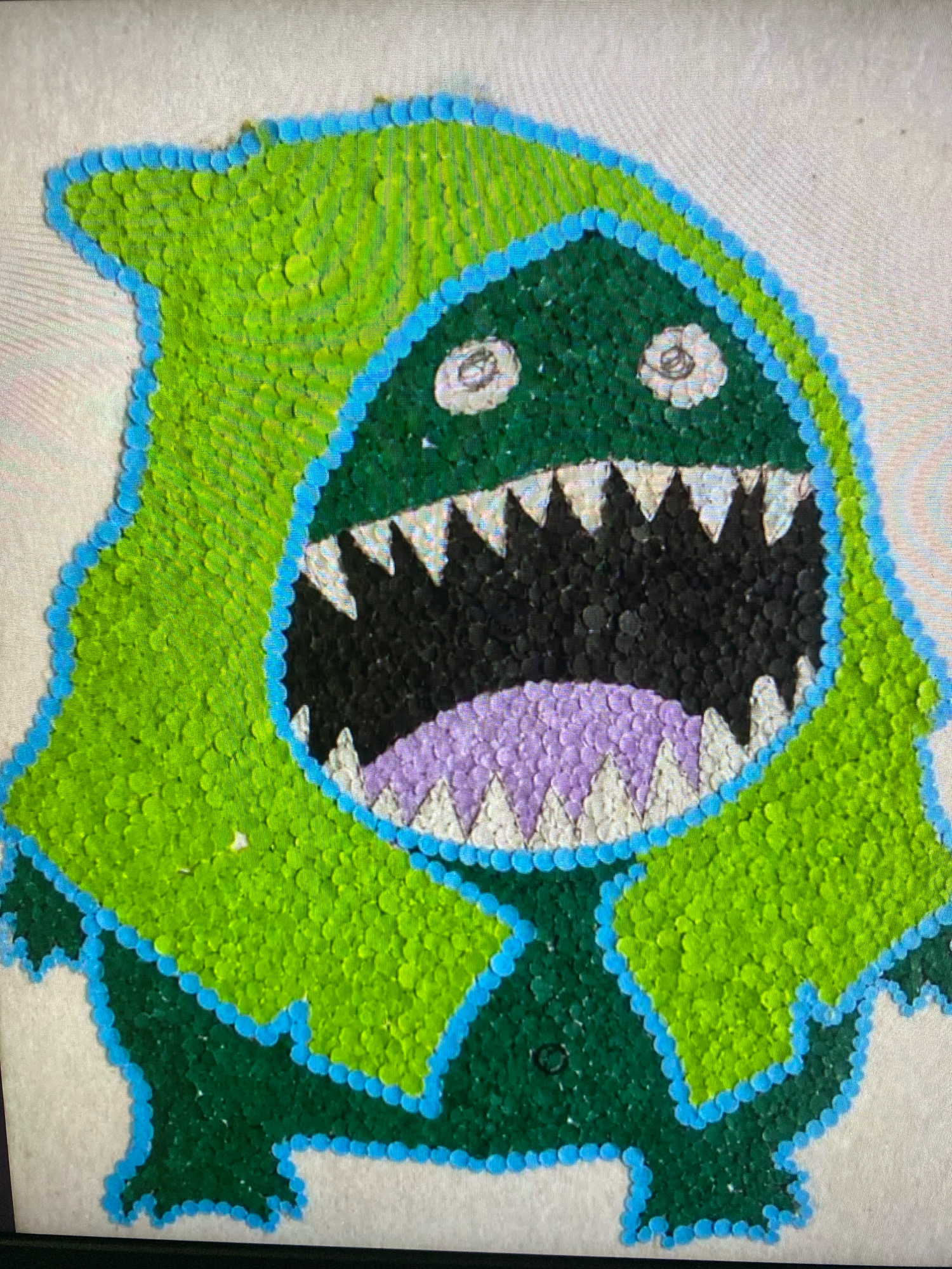The image shows a close-up, overhead view of a wall art. The art depicts a cartoonish green monster with a blue outline. The monster has a large, open mouth filled with black and purple sequins, and sharp white teeth. Its eyes are round and white with darker centers. The background appears to be a textured, light-colored surface. The image does not provide any information about its location in Cebu City, Philippines.