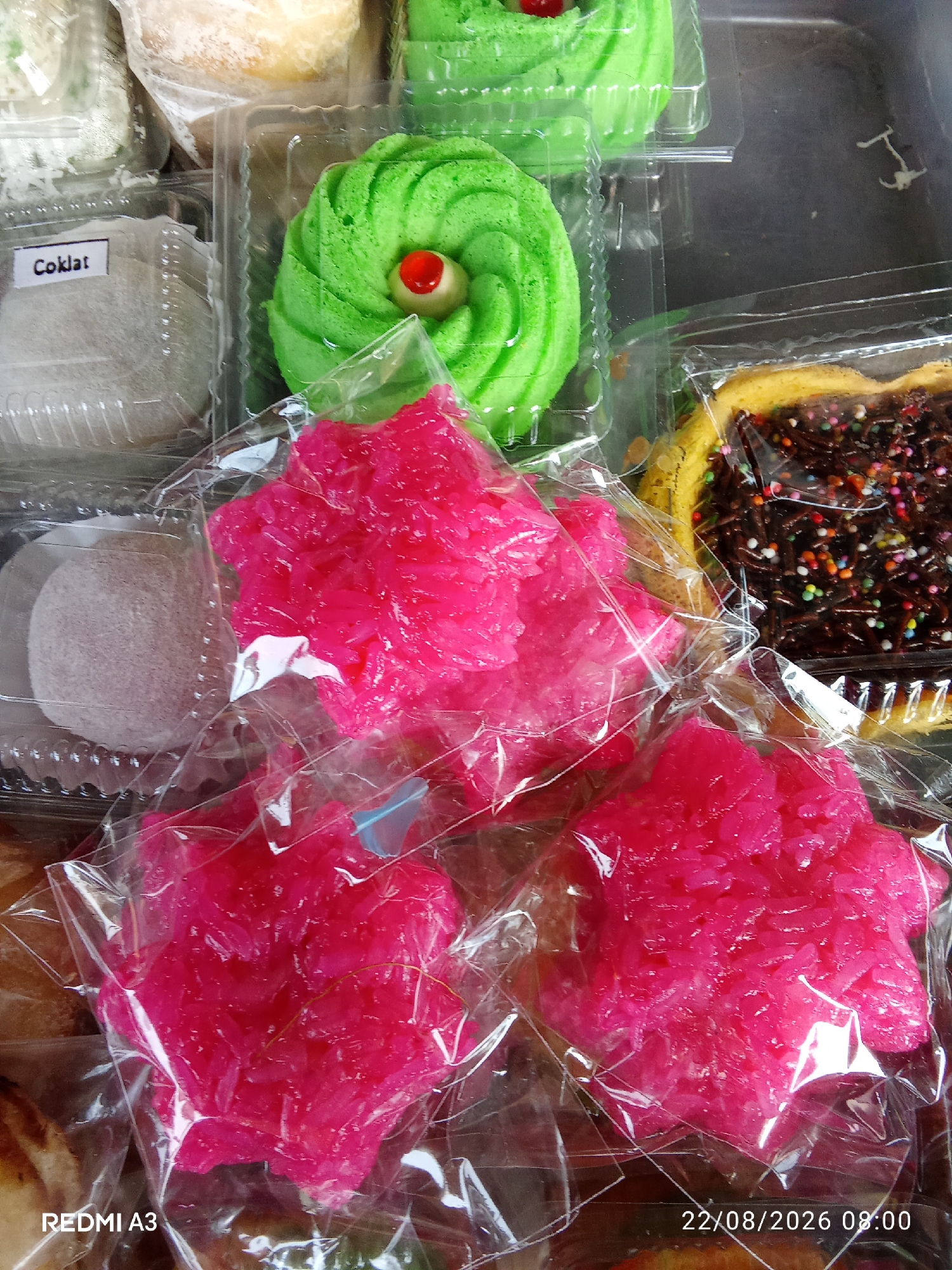 The image displays an assortment of colorful Indonesian sweets, likely a selection of Kue, arranged in clear plastic packaging. In the foreground, three vibrant pink, star-shaped sweets made of what appears to be shredded coconut and sugar are prominently featured, each individually wrapped in clear plastic. To the right of these, a slice of cake with chocolate frosting and rainbow sprinkles is visible. In the background, several green, swirl-shaped cakes topped with a red cherry and white cream are present, along with a white confection labeled "Coklat" (chocolate) in a separate container. The overall scene suggests a retail or market setting, possibly a street vendor or a small shop within Cemani, Indonesia, where such traditional sweets are commonly sold. The lighting is bright, indicating an outdoor or well-lit indoor environment. The image also includes a timestamp "22/08/2026 08:00" and the text "REDMI A3" in the bottom left corner, indicating the device and date the photograph was taken. There are no people visible in the image.