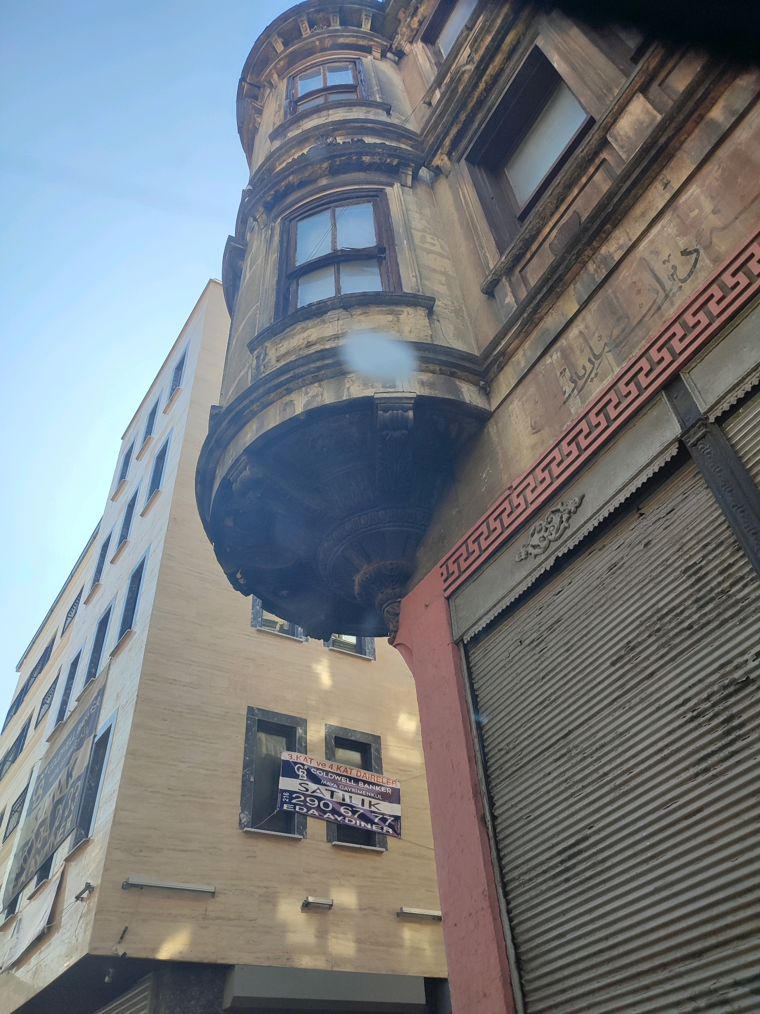 The image captures a low-angle view of two adjacent buildings in Istanbul, Turkey, under a clear blue sky. The building on the right is an older, ornate structure with weathered stone facade, featuring decorative Arabic script and a Greek key pattern border. It has a rounded bay window with dark wooden frames and a dark, elaborately carved overhang. Below this is a gray, corrugated metal roll-up shutter.
To the left, a taller, more modern building stands with a light-colored, textured facade and numerous dark-framed windows. A sign for "COLDWELL BANKER MAYA GAYRİMENKUL" is prominently displayed on this building, indicating a real estate agency. The sign, along with another partially visible poster on the left building, suggests a commercial area. The overall scene depicts a contrast between older, traditional architecture and newer construction in an urban setting.