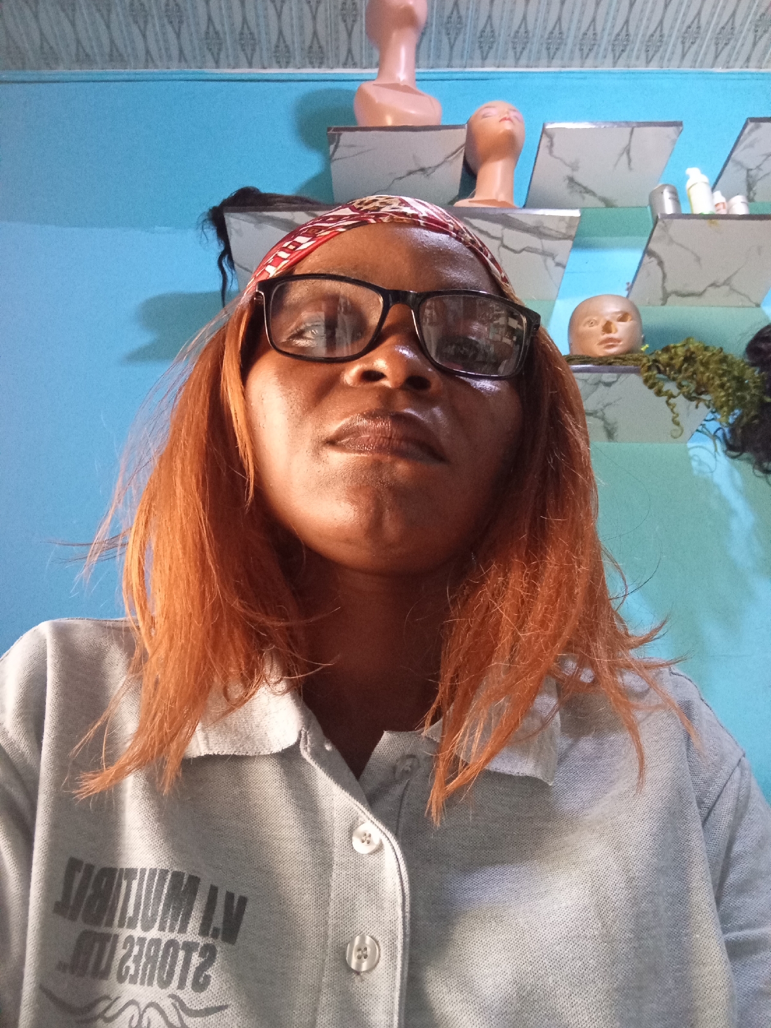 A close-up, low-angle, selfie-style photograph shows a person with reddish-brown, shoulder-length hair and fair skin wearing glasses. The person has on a light gray polo shirt with a collar and buttons, and the shirt has a dark, reversed text graphic on the front left side. The background features a bright blue wall, some shelves with mannequins and hair products, and decorative wallpaper at the top. The image appears to be taken indoors, possibly in a shop or salon. The reflections in the person's glasses suggest the presence of lights and possibly other shelves. The overall lighting is bright, creating some highlights on the person's face and clothing.