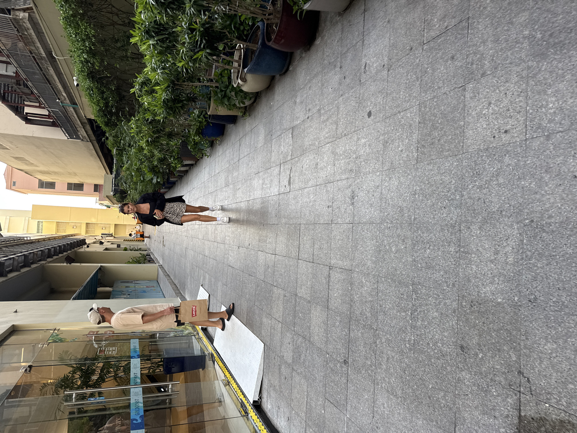 A man wearing a light-colored t-shirt, dark shorts, a baseball cap, and black sandals stands on a paved walkway, holding a brown paper shopping bag with the "Levi's" logo. Beside him, a woman in a black blazer over a patterned skirt and white sneakers walks towards the viewer. The walkway is lined with potted plants and leads into an alleyway between buildings. The man appears to be near the entrance of a building with large glass doors and signage that includes the words "GOLDCOAST." The background reveals an urban environment with buildings and more greenery. The overall scene depicts a street or a pedestrian area in a city. The context of Nha Trang Ward, Vietnam, is not directly discernible from the visual elements of the image alone.