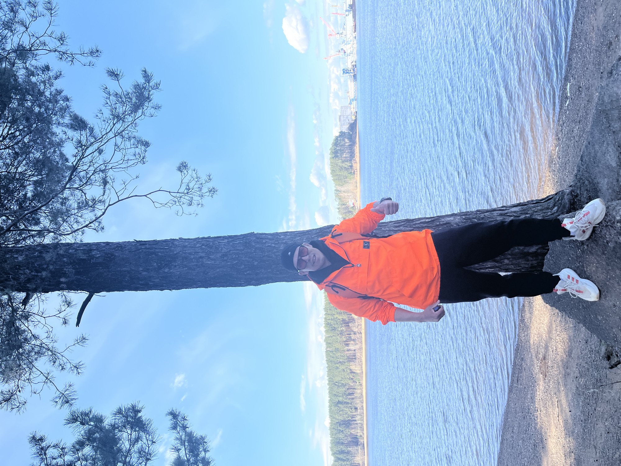 An outdoor scene captures a person posing against a large tree next to a body of water, likely in Chaikovsky, Russia. The image is originally rotated 90 degrees clockwise but depicts a vertical composition.
In the foreground, a person stands leaning against the sturdy, dark trunk of a conifer tree, which anchors the left side of the frame. The person is wearing a bright orange anorak-style jacket with a partial zip and a chest pocket, black pants, white athletic shoes with red stripes, a black beanie, and sunglasses. Their posture is relaxed, with one arm bent at the elbow.
To the right of the person and tree, a vast expanse of serene, blue water dominates the midground, reflecting the clear sky with gentle ripples on its surface. A distant treeline borders the far side of the water. Beyond this natural landscape, in the upper distance, an industrial area is visible with several cranes and buildings silhouetted against the horizon, suggesting a port or waterfront development.
The sky is a bright, clear blue with a few scattered white clouds, indicating a sunny day, likely morning or afternoon. The overall atmosphere is calm and bright, showcasing a blend of natural scenery and distant human infrastructure. There is no visible text in the image.