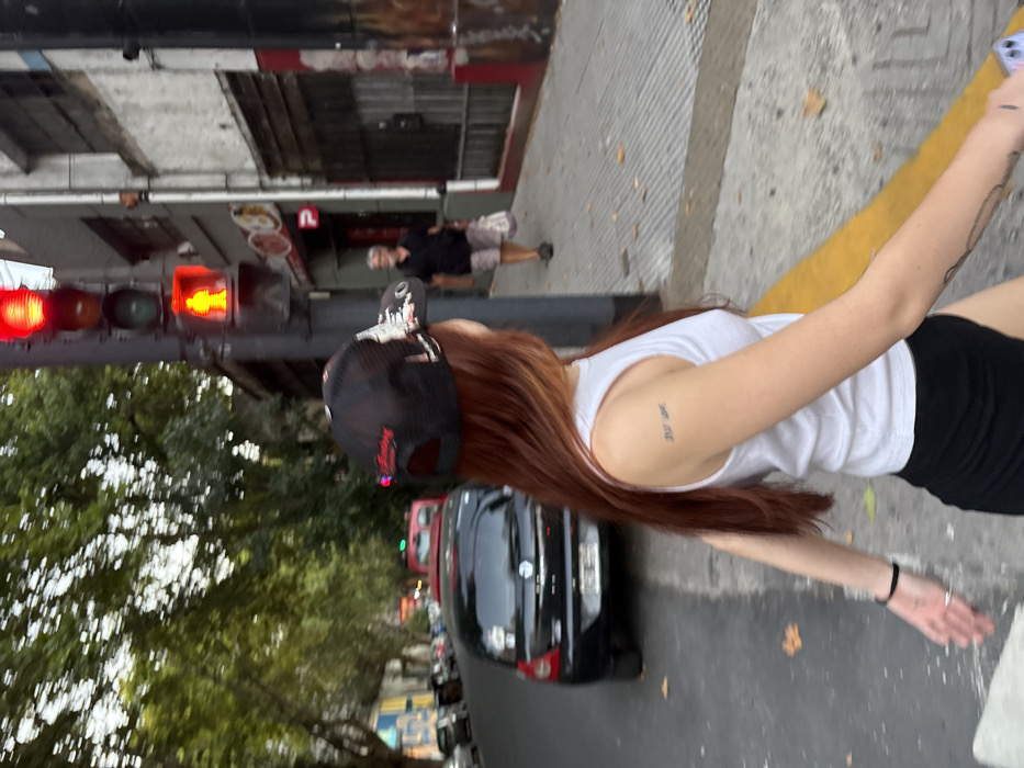 A young woman wearing a black baseball cap, white tank top, and black shorts stands on a city street. Her long auburn hair cascades down her back. A black car is parked behind her. A pedestrian crosswalk signal displays a red walking figure. Buildings line the street, with trees visible on the left. A yellow curb marks the edge of the sidewalk. Another pedestrian walks in the distance. The woman appears to be holding a smartphone.