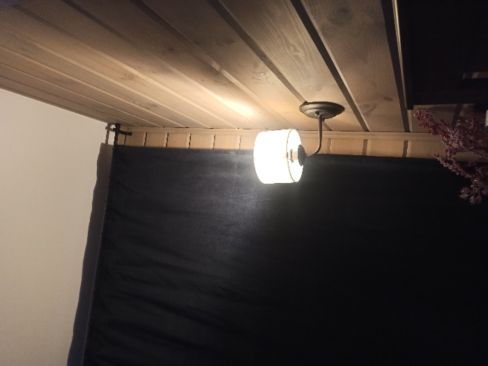 An interior scene located in Nizhneye Zadolgoye, Russia, depicts a room corner illuminated by a wall-mounted light fixture. The right wall is surfaced with horizontal light-toned wooden planks. Affixed to this wall is a sconce with a dark, curved metal arm supporting an opaque, cylindrical light shade that emits a warm, bright glow. To the left, a dark, solid-colored fabric curtain, likely black or deep navy, hangs from a dark rod positioned near the light-colored, flat ceiling. The curtain obscures the area behind it. In the lower right foreground, a dark, undefined object, possibly furniture, is partially visible, with a cluster of red or maroon artificial berries or flowers positioned atop or in front of it. No individuals are present in the scene, and no specific actions or interactions are discernible.