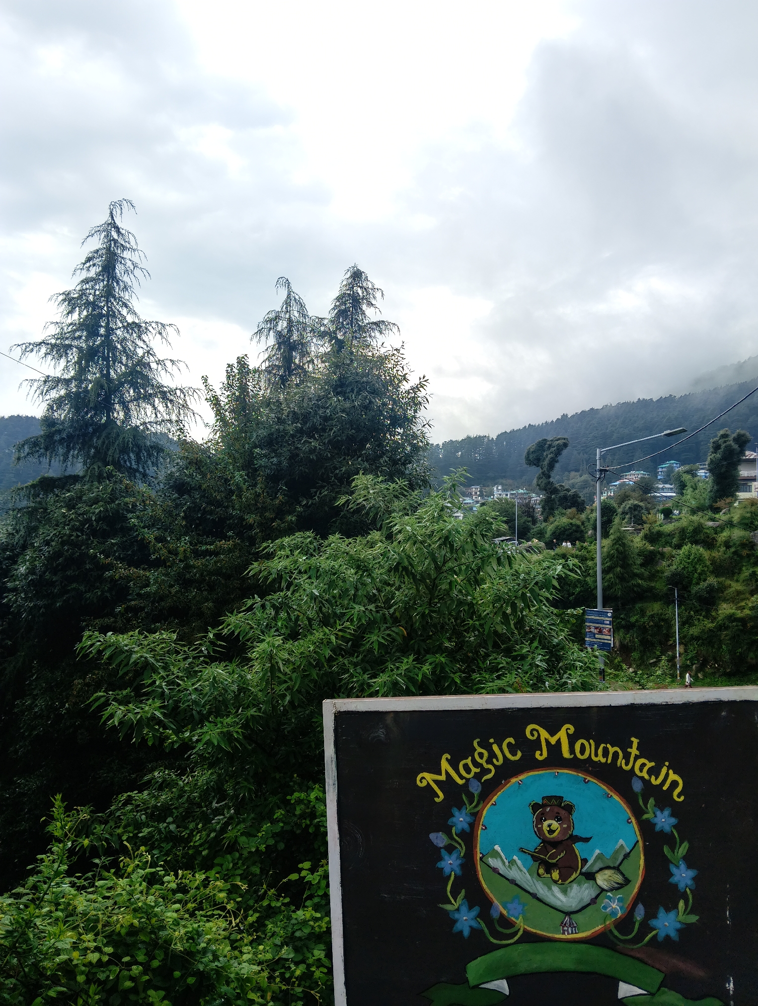The image depicts a mountainous landscape, likely in the city of Haini, India, with lush green trees and buildings scattered across the slopes. The sky is overcast, suggesting a cloudy or misty day. In the foreground, a black signboard features a colorful illustration. The illustration depicts a teddy bear wearing a hat and a scarf, riding a broomstick and flying over snow-capped mountains. Blue flowers with green leaves surround the central image. The signboard has a yellow inscription that reads "Magic Mountain" in a whimsical font. The overall impression is that of a scenic and somewhat mystical location, possibly a tourist attraction.