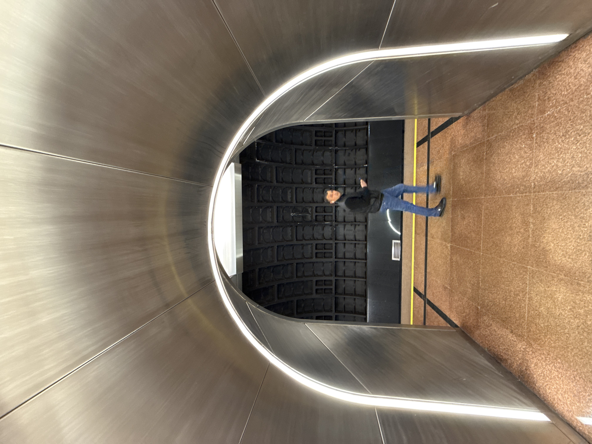 A person is standing in a tunnel-like hallway with a metal finish. The hallway curves around to the left, creating an archway effect with a light strip along the top. The person is wearing a dark jacket and blue jeans, and they are holding a phone in their hand. The floor of the hallway is a speckled brown color, and the walls on the left and right are different: the left side is the metal archway, and the right side is a textured surface. Behind the person, there is a black wall with recessed square patterns. The tunnel is located in the Zaryadye Park metro station in Moscow, Russia.