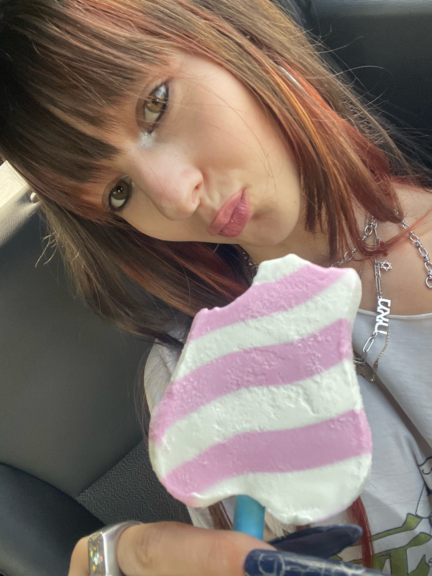 A young woman with brown hair and bangs, with streaks of red and orange, is holding a pink and white striped popsicle up to her face. She is wearing a white tank top and a silver necklace with Hebrew lettering and a Star of David pendant. Her fingernails are painted dark blue. She is wearing a ring on her left index finger. The background appears to be the interior of a car. The image was taken in Raanana, Israel.