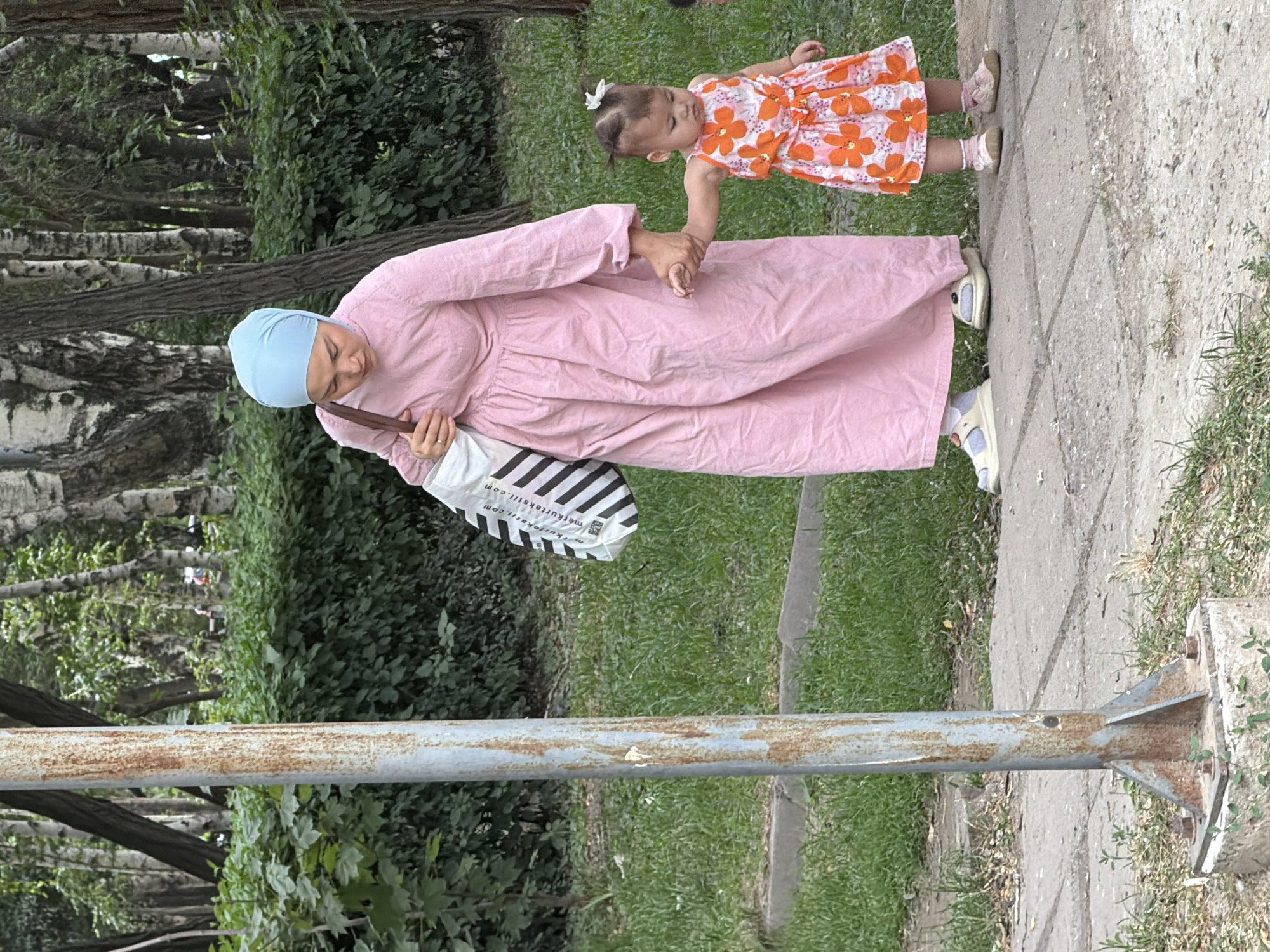 In an outdoor scene, a woman wearing a light blue hijab and a long, pink dress walks on a paved path beside a small child. The woman carries a large, black and white striped tote bag over her left shoulder. The child, wearing an orange and white floral dress, holds the woman's right hand. Both are walking from left to right in the frame. The environment consists of green grass and lush trees, with a rusty metal pole visible on the left side of the path. The overall setting appears to be a park or a treelined walkway in an urban area.