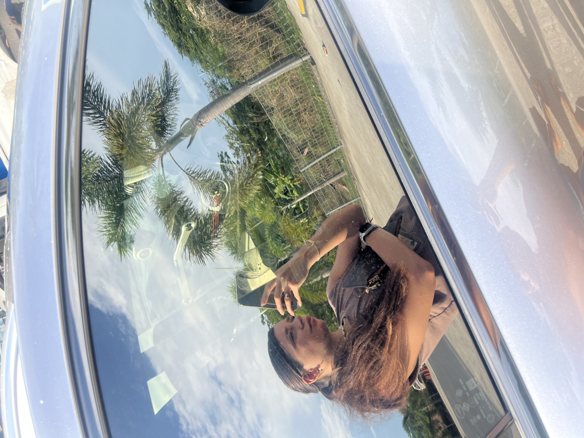 The image shows a reflection in the window of a car. A young woman is holding a smartphone and taking a selfie. The reflection also shows palm trees, a fence, and other vegetation. The location is unknown.