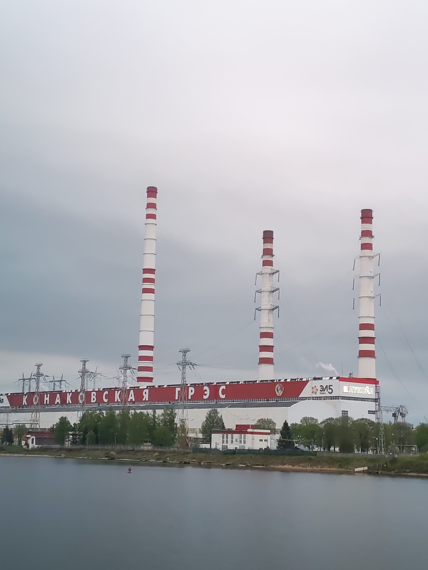 The image depicts the Konakovo Power Station, a large thermal power plant located in Konakovo, Russia. The plant features three tall smokestacks painted with alternating red and white bands. "КОНАКОВСКАЯ ГРЭС" is written across the top of the main building in large red Cyrillic letters. The power station is situated on the bank of a body of water, presumably a river or lake. Power lines and transmission towers are visible, carrying electricity away from the plant. The sky is overcast, indicating potentially inclement weather. Trees and vegetation are present along the shoreline.