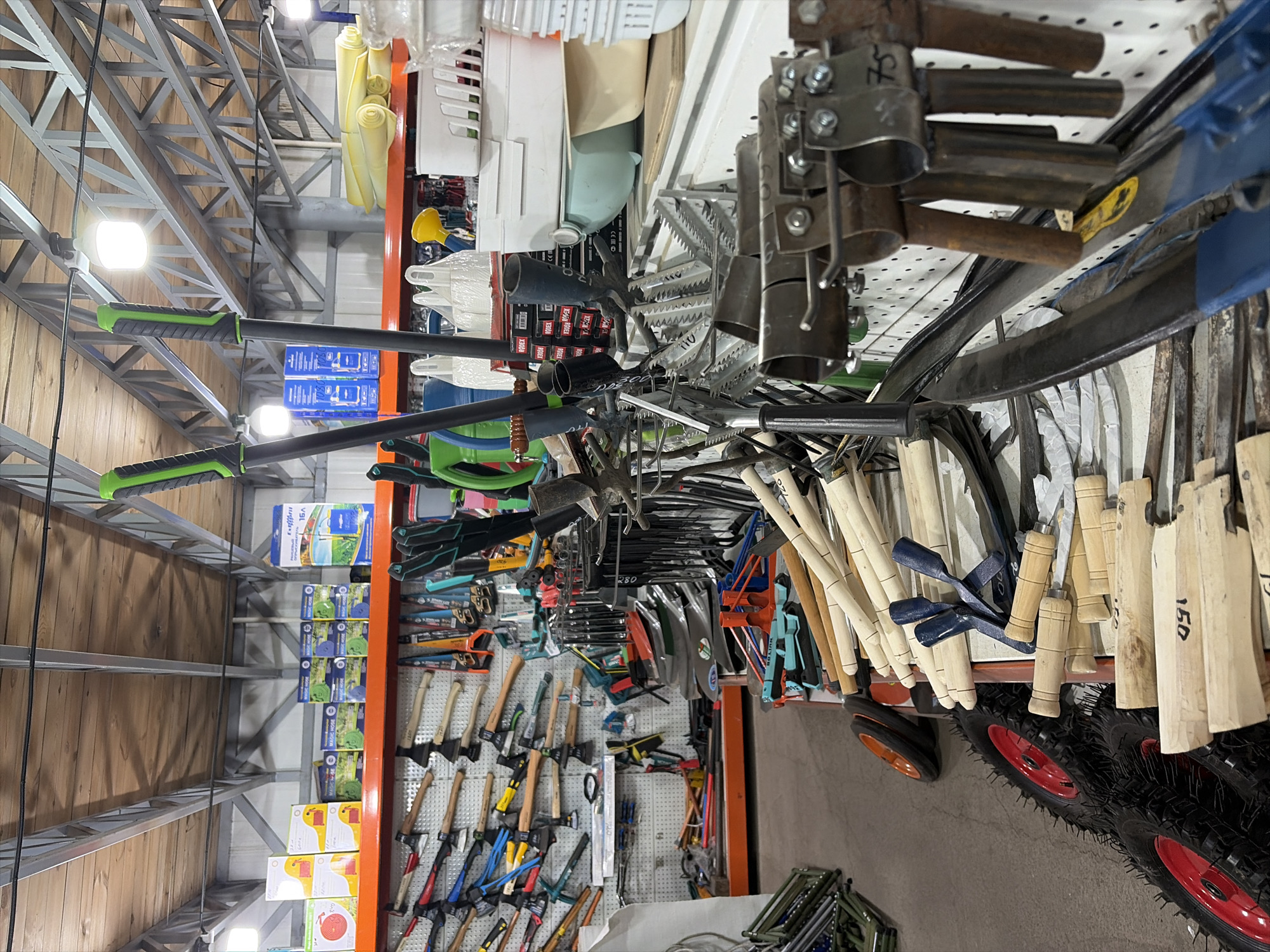 The image shows a retail store interior in Kant, Kyrgyzstan, specializing in gardening tools. The store is densely stocked with various items displayed on shelves and hung on pegboards. In the foreground, there is a collection of wooden-handled tools such as trowels, small shovels, and what appear to be parts of gardening hoes. To the left, a stack of large, spiky tires with red rims are visible. Further back, on a shelf, are numerous axes and hatchets with wooden handles hanging on a pegboard. Above, on higher shelves, are boxes of gardening hoses and other packaged gardening supplies. Various metal tools, including rakes and pruning shears, are also displayed. The overall environment appears to be a well-organized, albeit crowded, retail space. There are no people visible in the image.