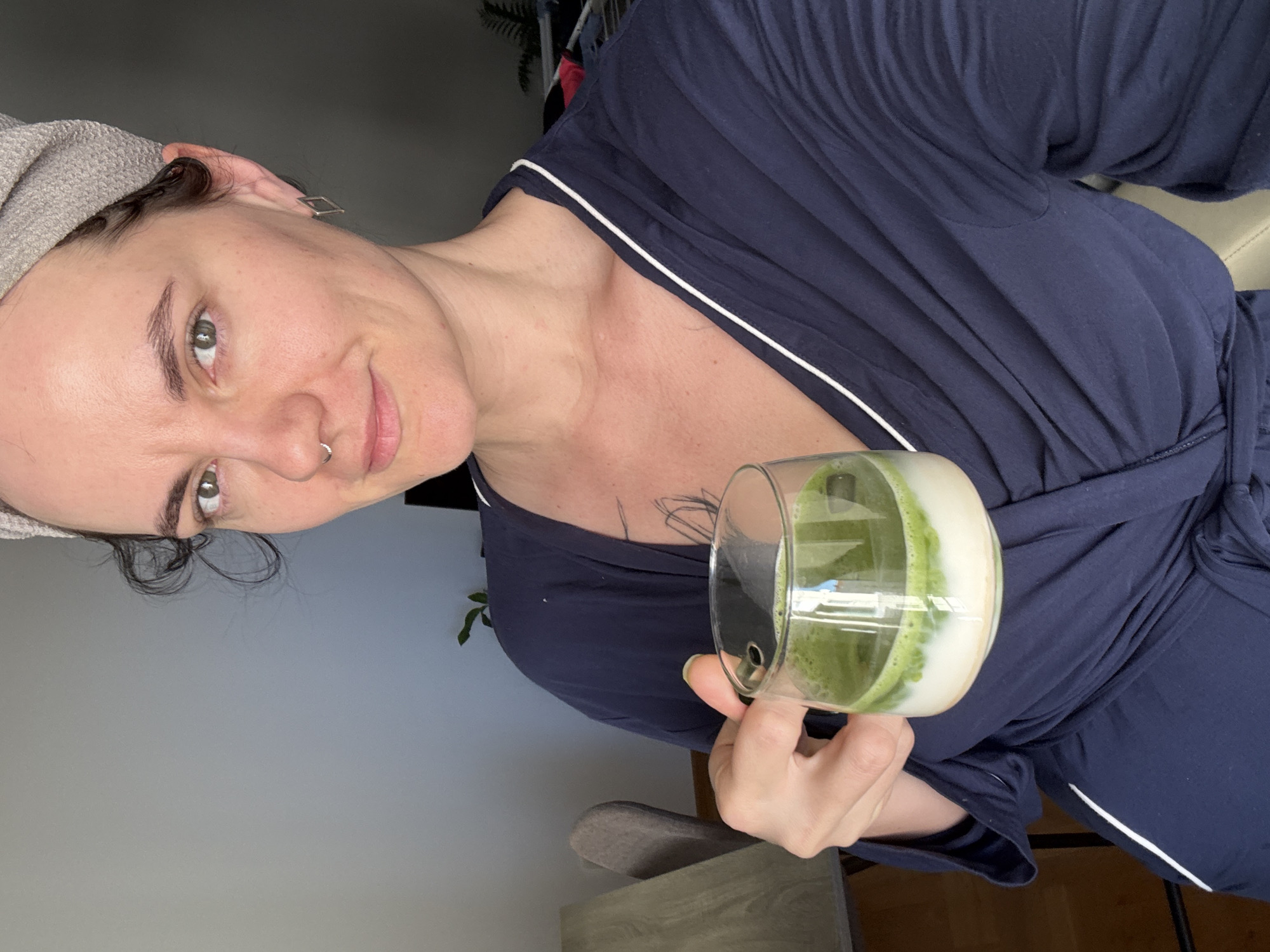 A person is holding a glass of matcha latte. The person has light skin and is wearing a dark blue robe with a white trim. They have a silver band in their hair and a nose ring. The glass contains a green and white liquid, indicating matcha latte. There is a plant behind the person. The location is not discernible from the image.