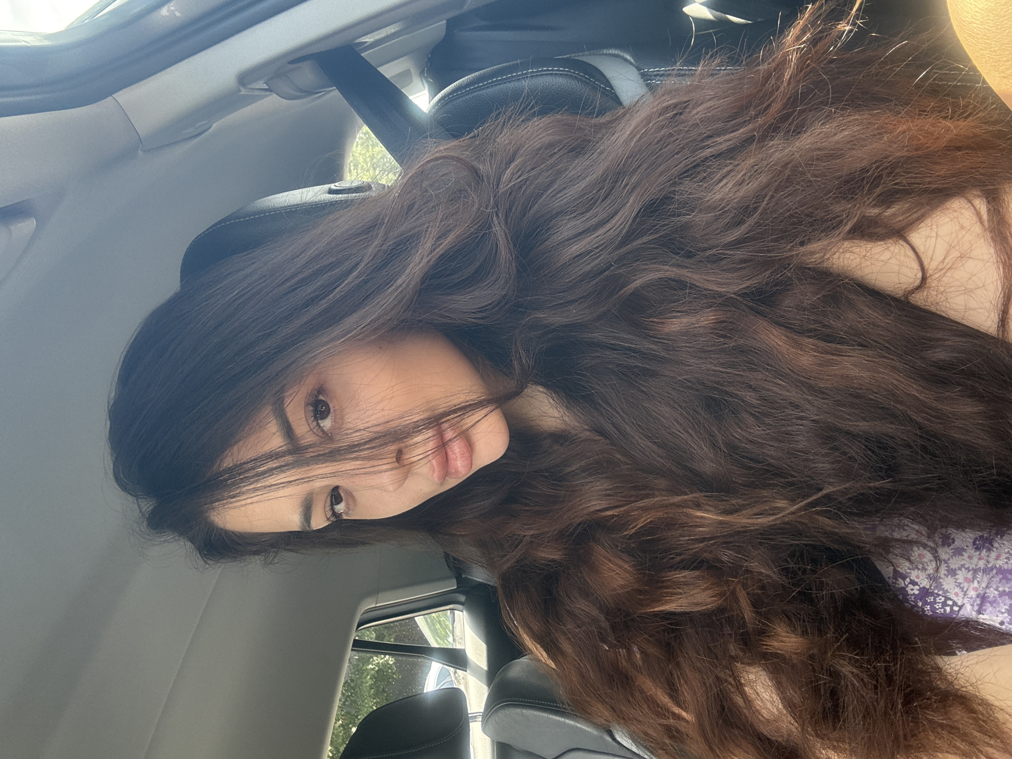 A woman is seated in a car. Her long, wavy brown hair is partially obscuring her face. She is wearing a floral-patterned top, with hues of purple and white. The car interior is visible. The background shows a partial view of a window. The location cannot be determined from the image.