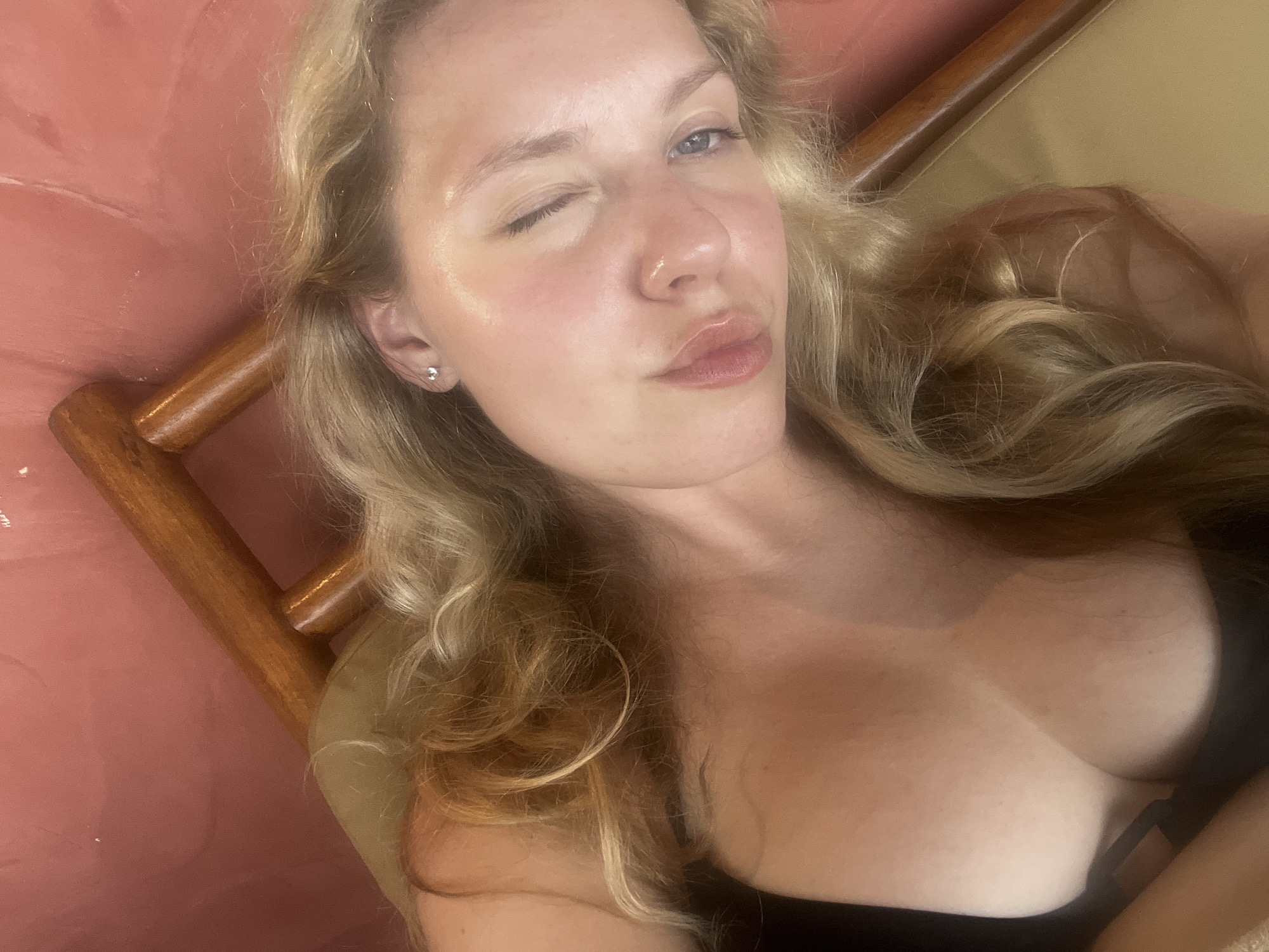 A woman with light blonde hair and fair skin is lying down, possibly on a lounge chair. She is wearing a black top that reveals cleavage. Her head is tilted towards the camera, and she is winking with her left eye while pursing her lips in a playful manner. She is wearing a small earring in her left ear. The background appears to be a textured surface, possibly a wall or part of the furniture, with hints of pink and a wooden frame around her. The image appears to have been taken indoors or in a sheltered area, perhaps a resort or private residence. The city is assumed to be Mengwi, Indonesia based on the prompt request.