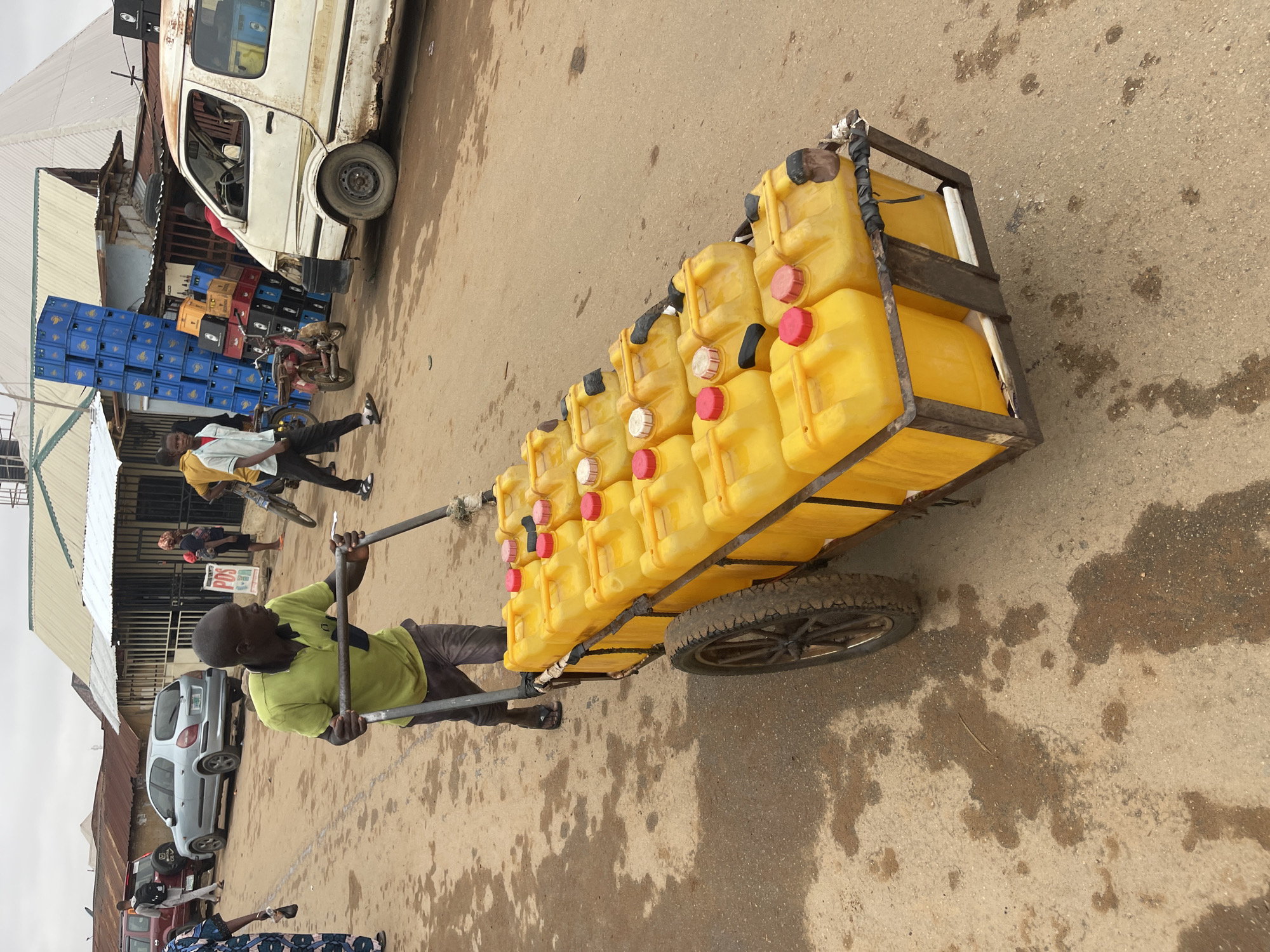 A man is pulling a yellow cart laden with yellow containers, possibly jerrycans, down a street in Kuchingoro, Nigeria. The cart has a metal frame and two wheels. The man is wearing a light green shirt and dark-colored trousers. The street surface appears to be concrete or asphalt with stains and discoloration. In the background, there is a white van, blue crates stacked, a building with a corrugated metal roof, and other people. A silver car is parked along the side of the street.