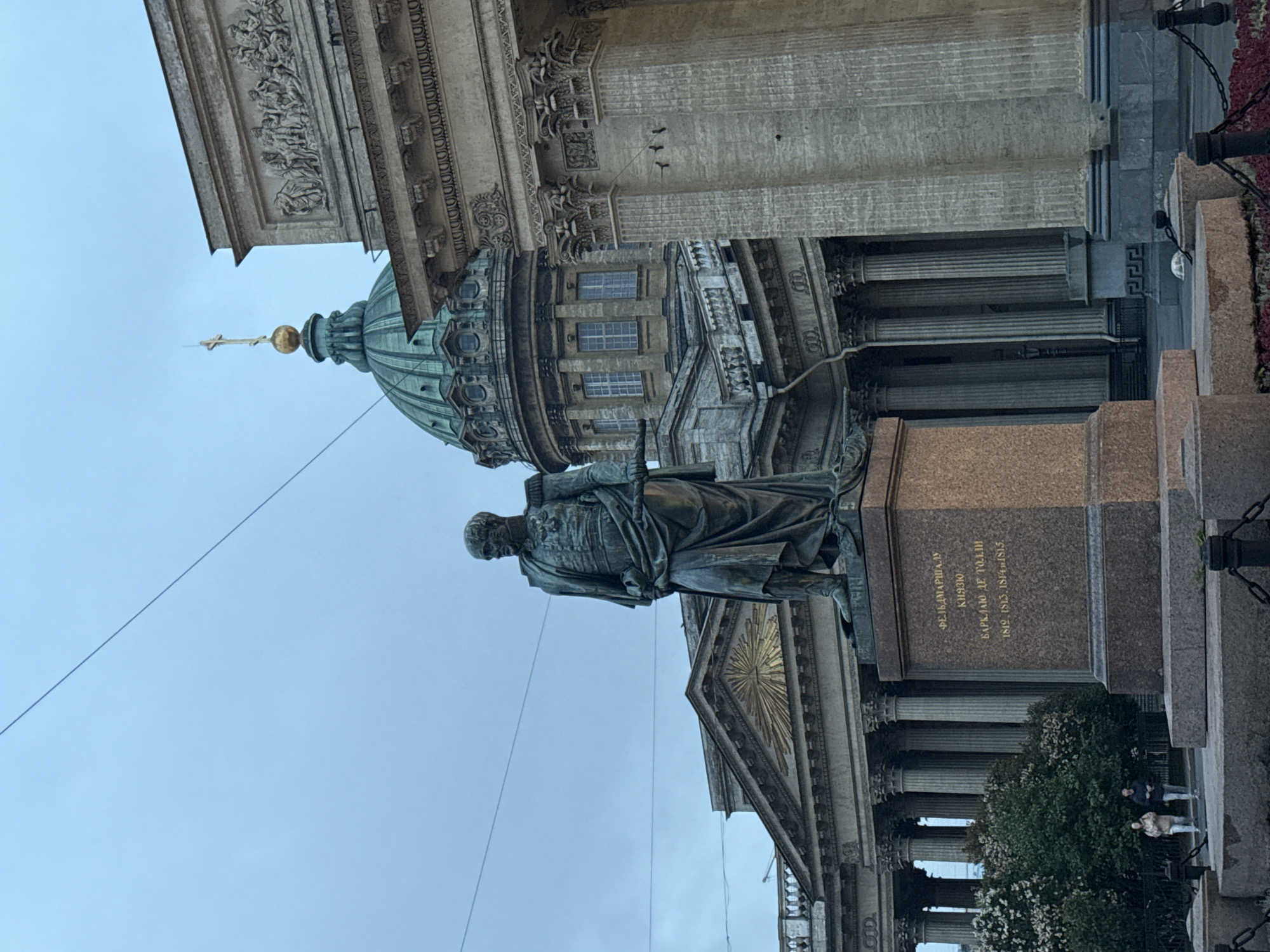 This is a medium shot, exterior, eye-level photograph of a bronze statue on a granite pedestal in front of the Kazan Cathedral in Saint Petersburg, Russia. The sky is overcast and a pale blue. The statue depicts a man in military uniform, with a cloak draped over his shoulders. The pedestal is inscribed with Russian text. Behind the statue, the Kazan Cathedral's semicircular colonnade and green dome are visible. The architecture is neoclassical. Two people, dressed in casual clothing, are walking in the lower left portion of the frame, near some green bushes. A few power lines are visible in the upper portion of the frame.