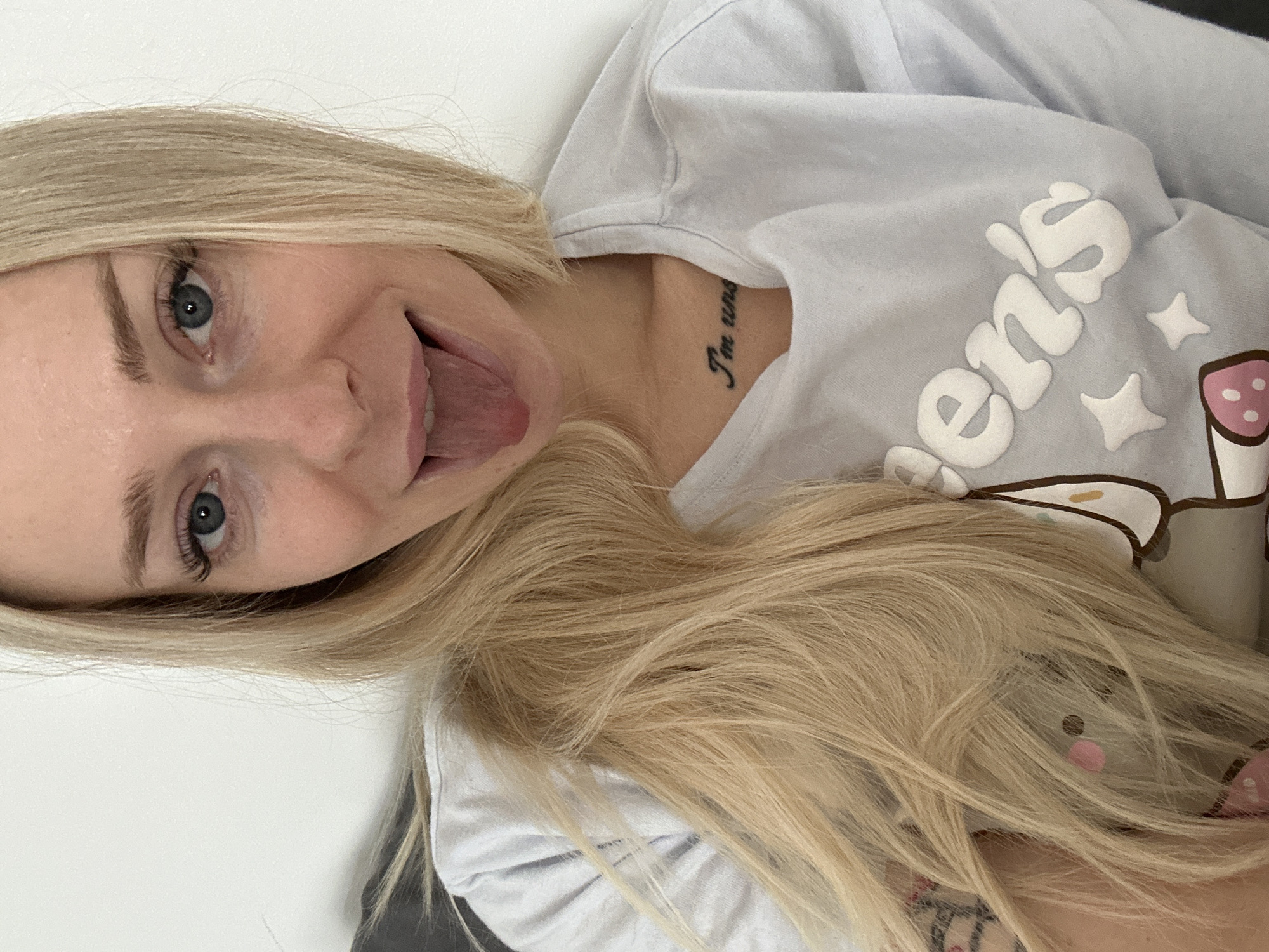A close-up, head-and-shoulders shot of a woman with blonde hair and blue eyes is shown against a plain white wall. She is wearing a light blue t-shirt with white lettering that says "Queen's" and some cartoon characters. A tattoo is visible on her chest. The woman is sticking her tongue out. The image does not contain any information about the city of Krakow, Poland, or any specific environmental details that would place it there.