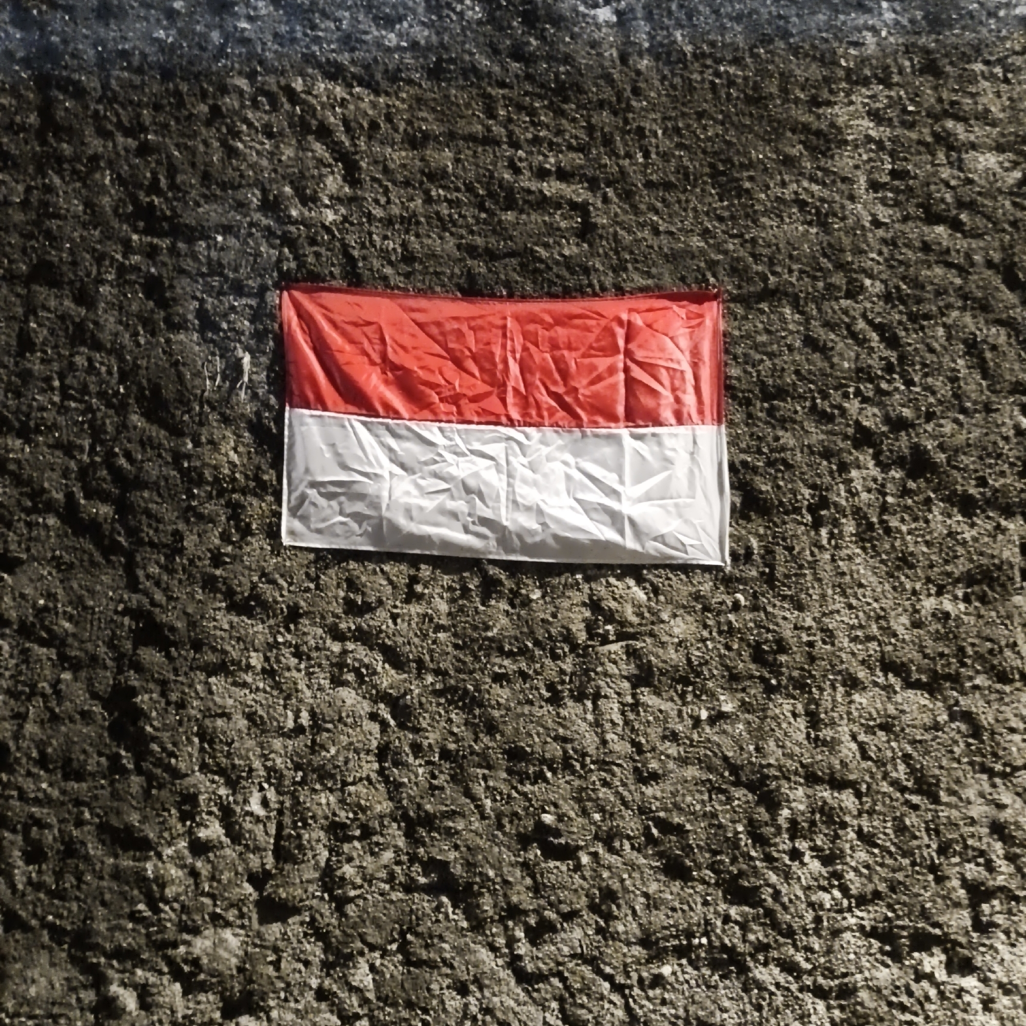A red and white flag is attached to a rough, dark gray textured wall. The flag is positioned slightly off-center to the left, horizontally, with the red stripe on top and the white stripe below. The flag appears to be made of fabric and has visible creases. The wall has a granular, uneven surface with small indentations and protrusions. The lighting suggests the image was taken outdoors, possibly during the day, with a shadow cast on the upper left portion of the wall. There is no indication of any specific location within South Jakarta, Indonesia.