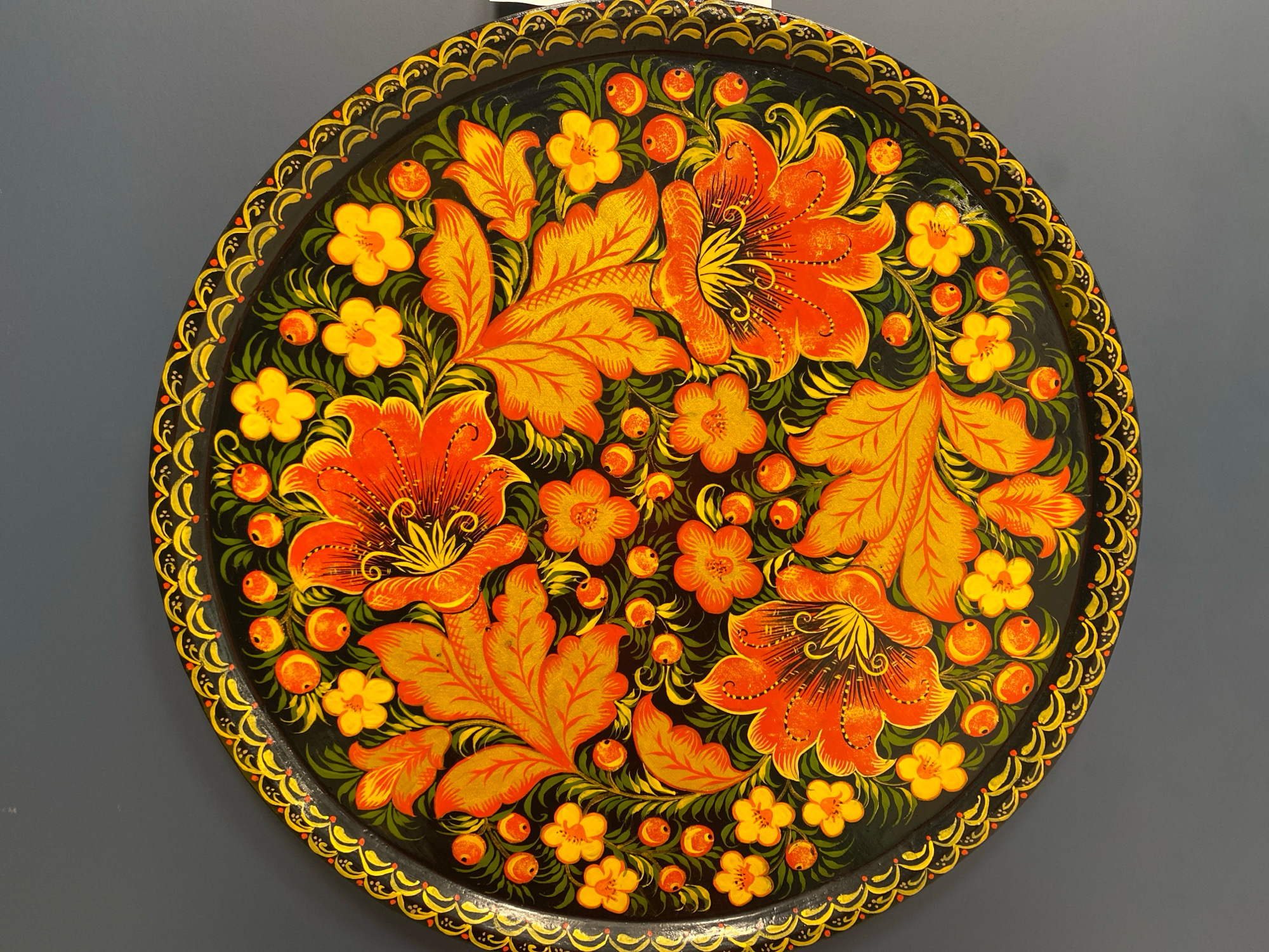 The image shows a round, black metal tray adorned with vibrant, hand-painted floral motifs. The main design features large, stylized orange and yellow flowers with intricate details, interspersed with smaller yellow flowers and clusters of small orange berries. Green foliage and delicate tendrils fill the spaces between the flowers. The rim of the tray is decorated with a patterned border in yellow and red. The tray is displayed against a plain, dark gray background. There is no discernible information about its specific location in Ivanteyevka, Russia, within the provided image.