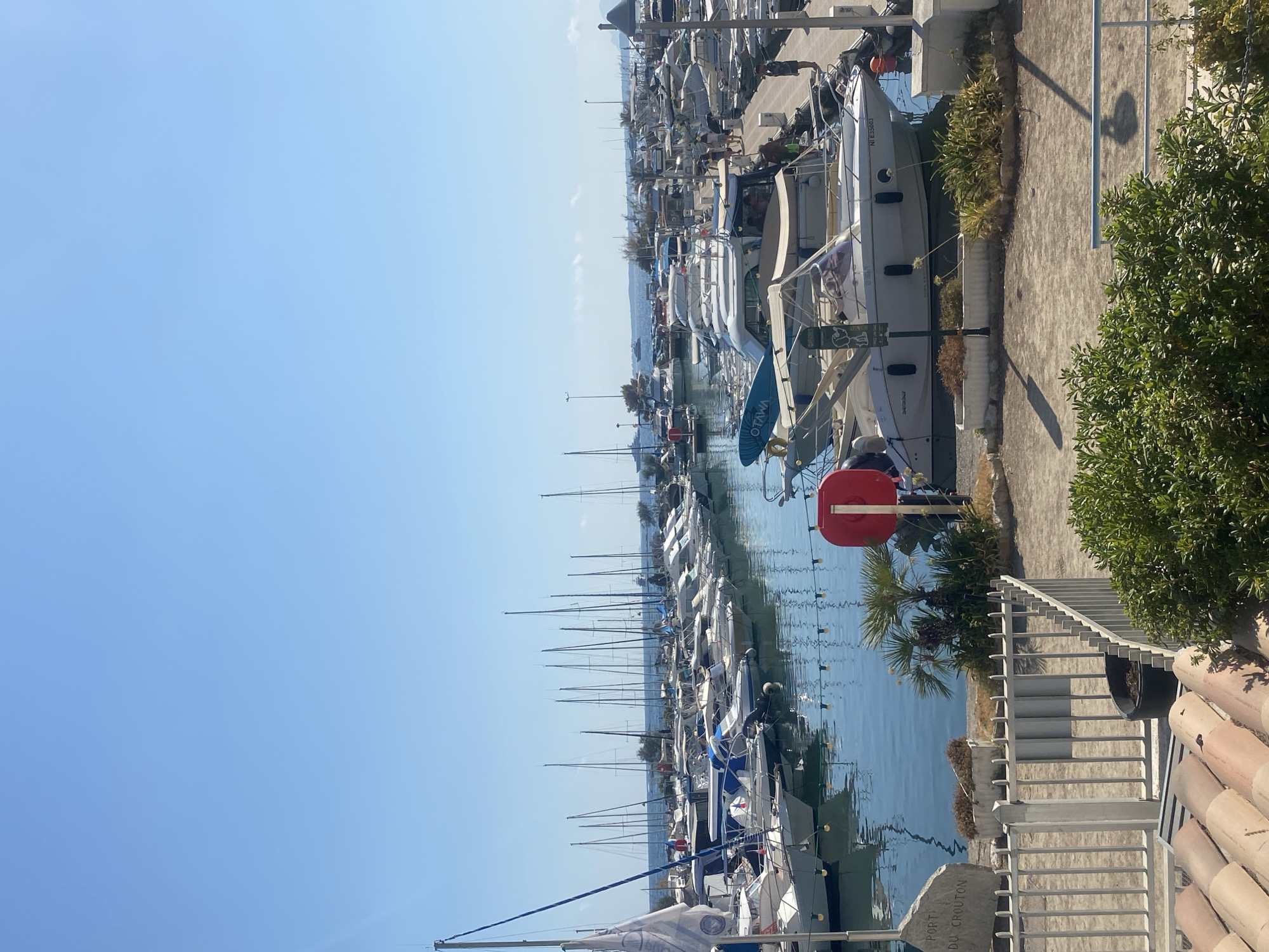 The image depicts a sunny day at the Port du Crouton in Antibes, France, a harbor filled with numerous docked boats of various sizes, primarily motorboats and sailboats. The water in the harbor is calm and reflects the clear blue sky. On the left side of the image, a large stone monument reads "PORT DU CROUTON," confirming the location. A white railing and some landscaping, including palm trees and bushes, are visible along the waterfront. Several people are present on the docks, some walking and others seemingly relaxing. The boats are tightly packed, suggesting a busy marina. The overall scene is one of a tranquil day at a Mediterranean port.