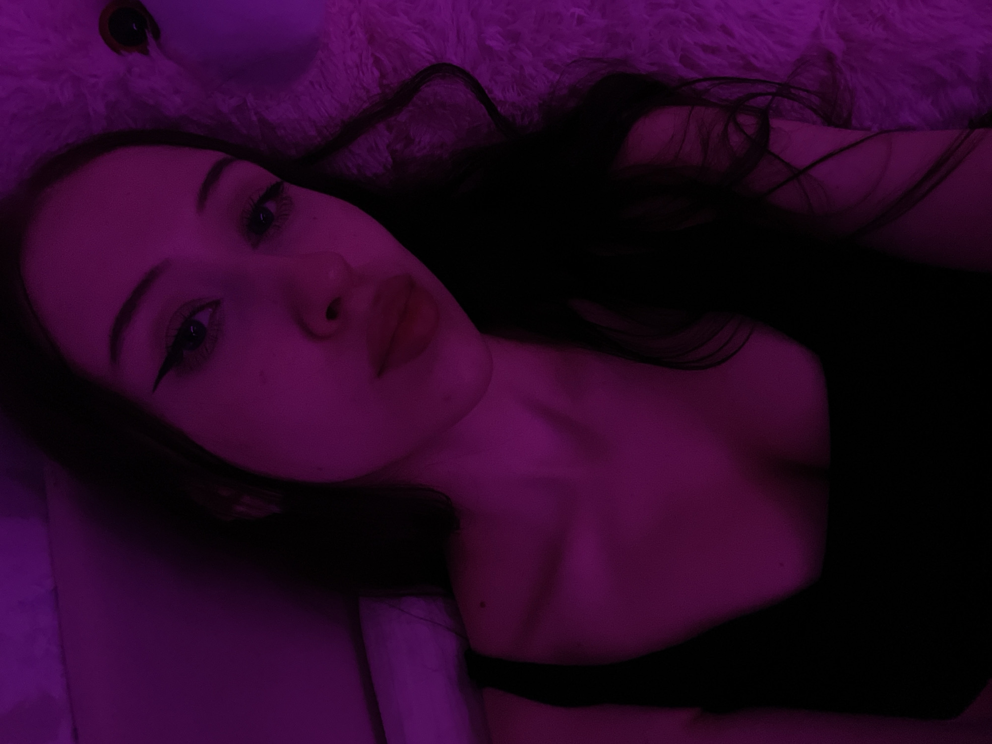 The image shows a close-up, waist-up portrait of a young woman with dark hair, bathed in purple lighting. She is wearing a black tank top. Her makeup includes eyeliner and mascara. The woman is lying down on a fluffy, textured surface. The background is indistinct and also illuminated by purple light, suggesting an indoor setting. There is no discernible connection to the city of Votkinsk, Russia.