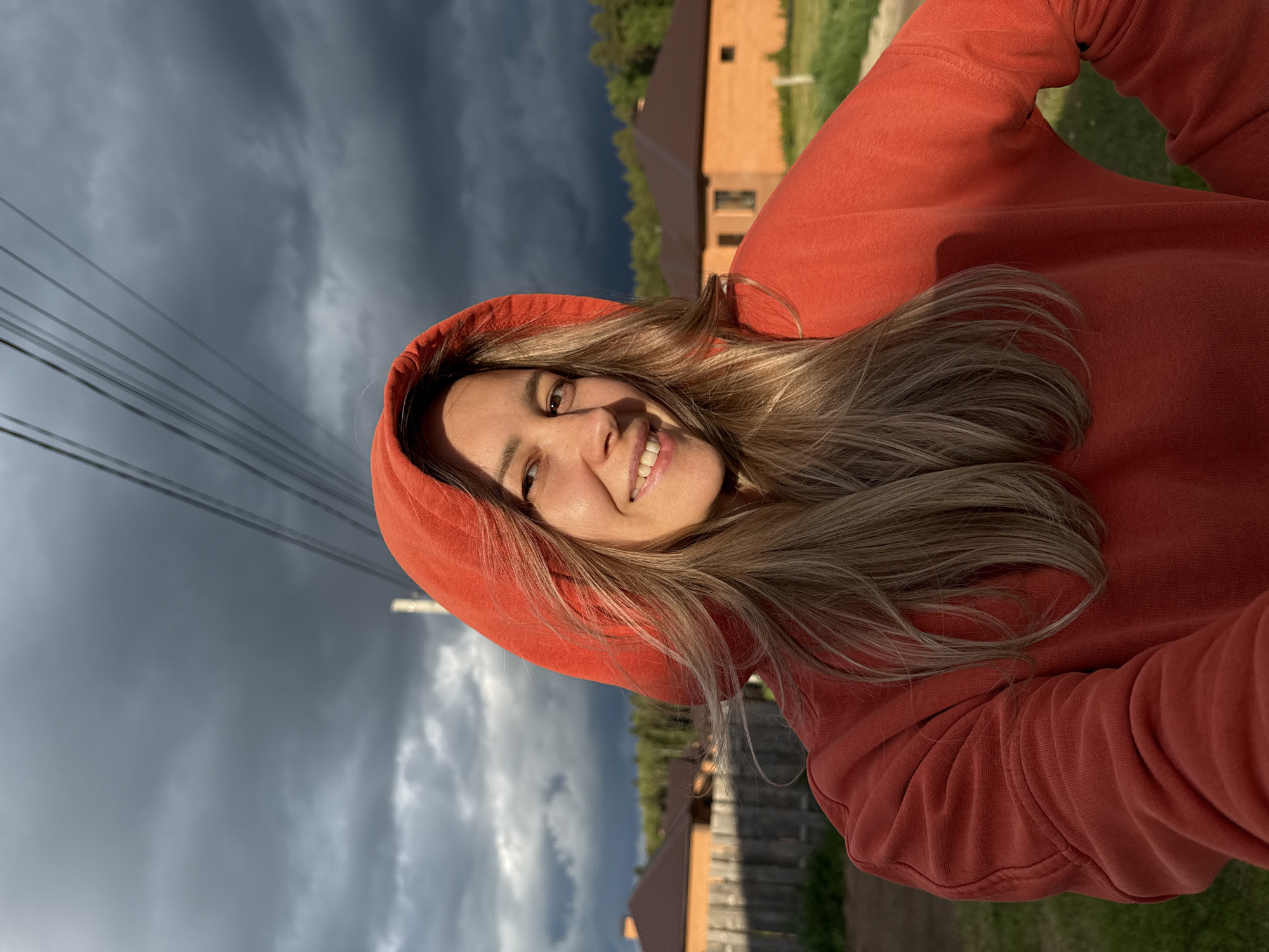 A woman with light brown hair, wearing an orange hoodie, is smiling at the camera. She appears to be outdoors. Overcast skies are visible behind her. A building with a reddish-brown roof is in the background. Power lines run across the upper left portion of the image. The scene is set in a residential area, potentially the city of Chaikovsky, Russia.