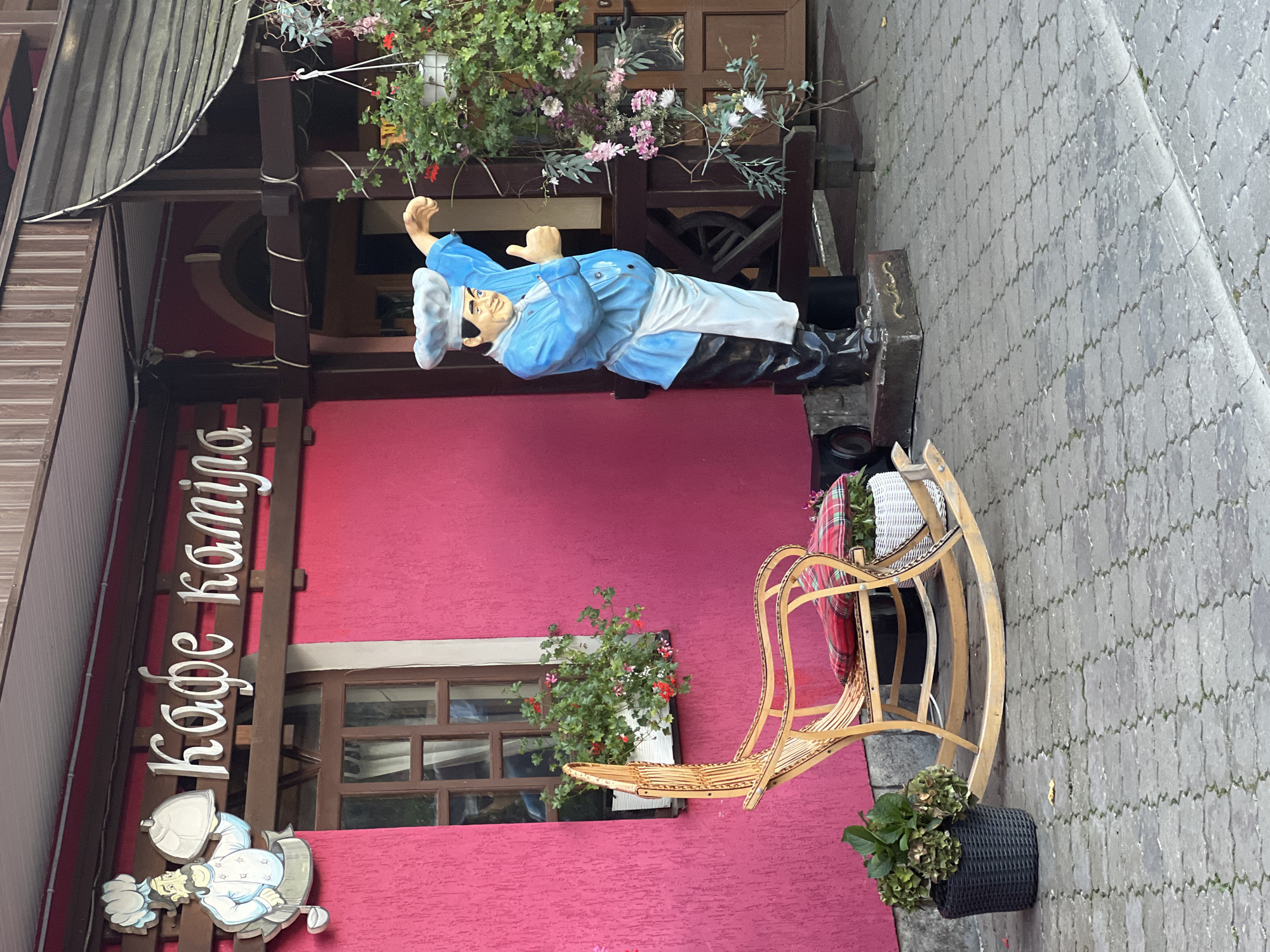 The image depicts the exterior of a restaurant named "Кафе Каміла" (Cafe Kamila) in the city of Сонячне, Ukraine. A bright pink stucco wall serves as the backdrop, with a wooden-framed window adorned with flower boxes containing red and green plants. In front of the window, a light-colored wooden rocking chair with red plaid cushions is placed on a paved walkway made of interlocking gray stones.
To the right of the chair, a life-sized statue of a chef stands, wearing a blue uniform, a white apron, and a tall chef's hat. The statue is posed with one arm extended and a thumb up, as if giving approval. Hanging plants with pink and white flowers are visible to the right of the statue, alongside a wooden railing and a brown wooden door.
A cutout sign above the window features a cartoon chef holding a covered dish, next to the restaurant's name in white lettering. The overall scene suggests a welcoming and somewhat whimsical atmosphere for a cafe.