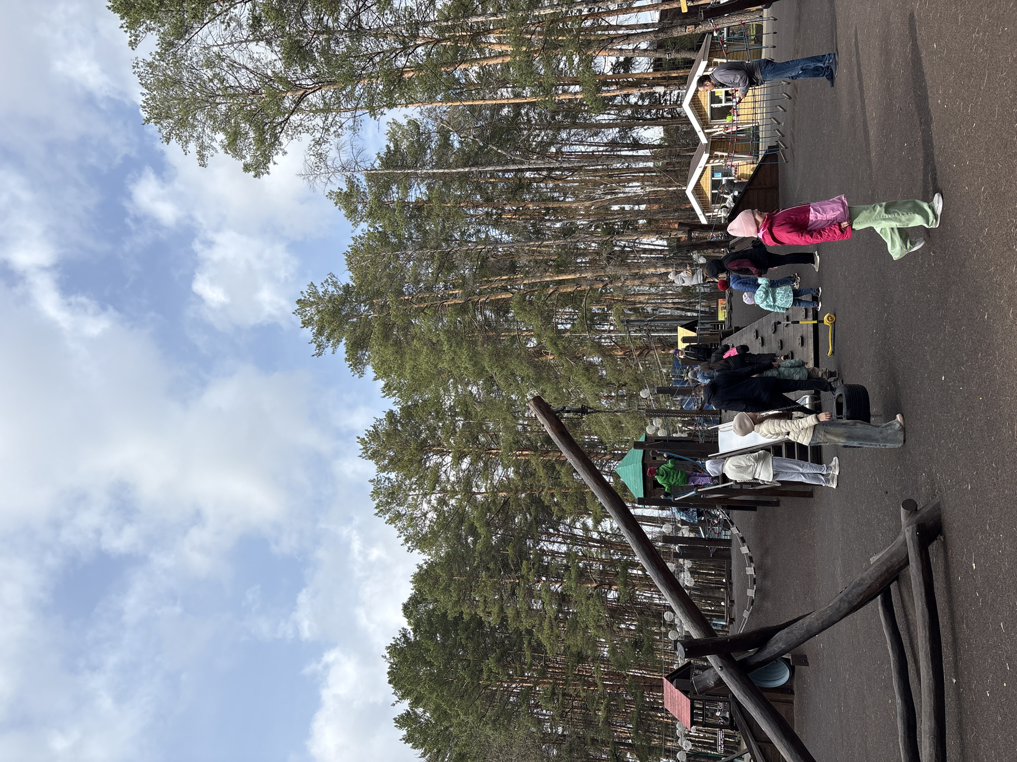 The image shows a playground or outdoor recreation area in Neftekamsk, Russia. A group of people, including children and adults, are participating in activities at the playground. Tall pine trees surround the area, with the sky partially visible. The playground equipment appears to be made of wood and rope. There are small wooden structures resembling houses or play areas. Some individuals are interacting with the equipment, such as climbing or balancing, while others are standing or observing.