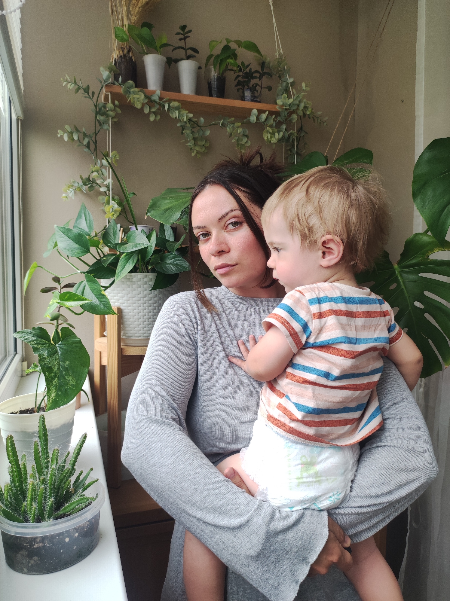 A woman with dark hair is holding a toddler with blonde hair in her arms. The woman is wearing a long-sleeved grey shirt or dress, and the toddler is wearing a striped shirt and a diaper. The woman has a serious expression on her face, and the toddler is looking to the side.
The background consists of a wall with several plants on shelves and near the window. There are different types of plants, including succulents, leafy plants, and some plants on a wooden shelf. A window is visible on the left side of the frame. The setting appears to be an indoor space with ample natural light.
There is no location data provided.