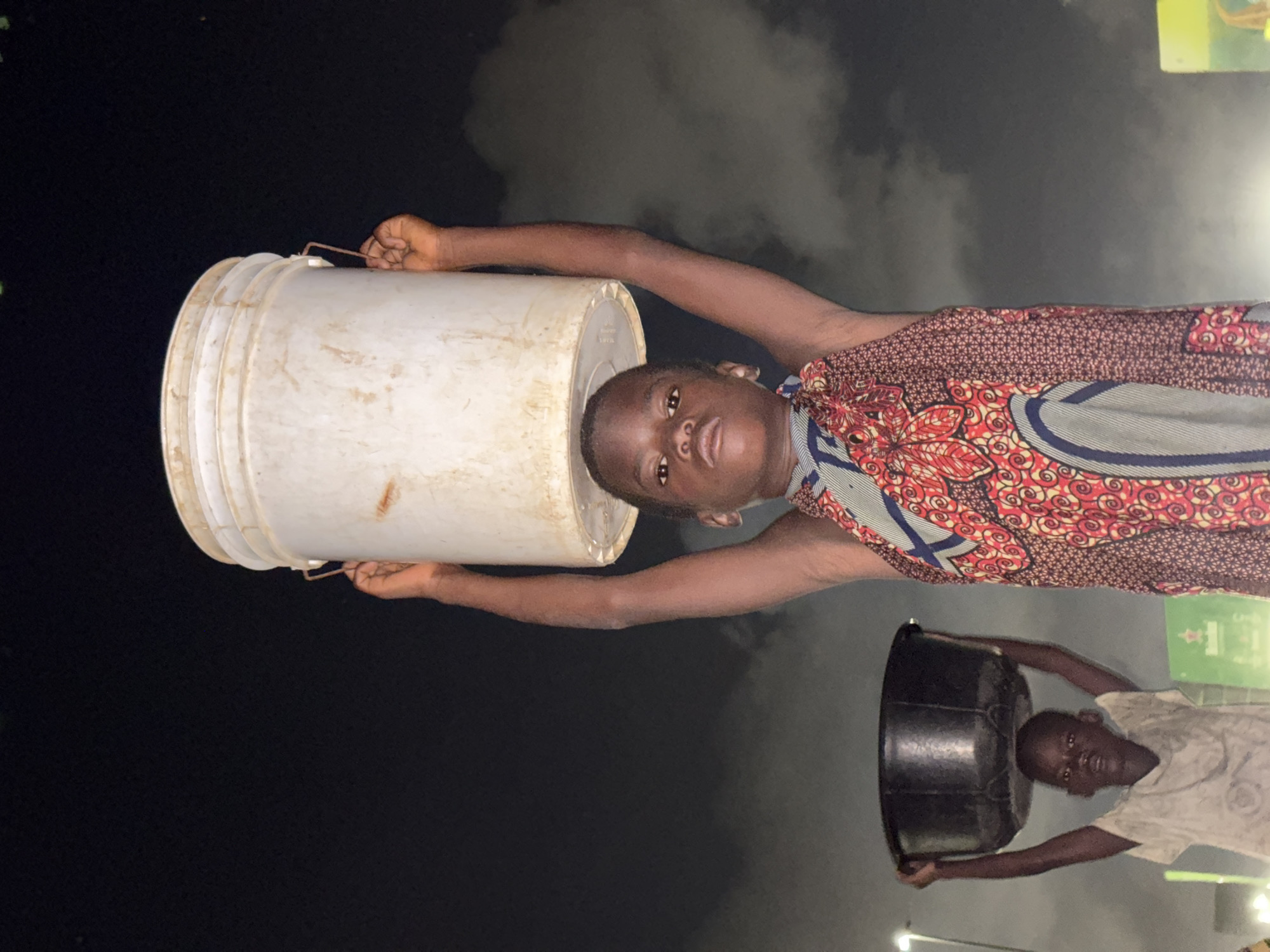 A young person is carrying a large white bucket on their head. The person is wearing a patterned shirt. Another person carries a black bucket on their head, and they are wearing a light-colored shirt. The background is dark. The location and specific actions are not able to be determined from the image.