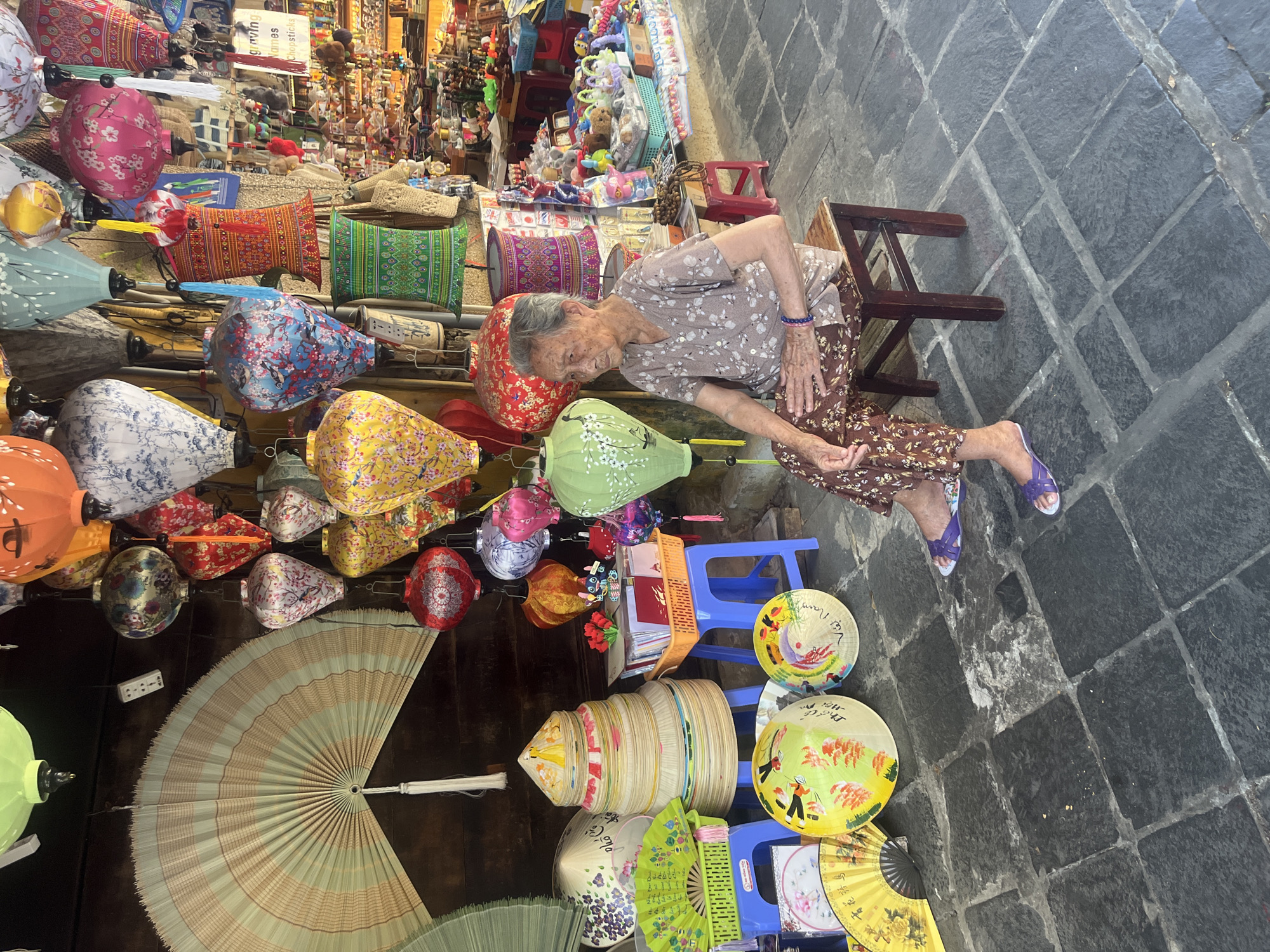 This media file captures a vibrant street market scene in Hội An Ward, Vietnam, showcasing a variety of local crafts. The image is oriented vertically, depicting an elderly woman seated on a wooden stool on the right side of the frame. She has grey hair, wears a floral-patterned light brown top and matching long pants, and purple sandals. A blue bracelet adorns her left wrist. Her posture suggests a moment of rest or quiet observation.
Around her and filling the left and foreground are numerous colorful handmade items. Prominently displayed are various silk lanterns in different shapes (round, elongated, cylindrical) and patterns, including floral, geometric, and landscape designs, in shades of red, pink, blue, yellow, green, orange, and white. Traditional Vietnamese conical hats (Nón Lá) are stacked and arranged on blue plastic stools, some plain, others beautifully hand-painted with scenes of people and nature. Hand fans, both large and small, with intricate designs, are also visible.
In the background, shelves and stalls overflow with other merchandise, including brightly patterned pillows, woven baskets, and small souvenirs such as plush toys and keychains. A sign partially reads "Engraving Names Chopsticks," suggesting customization services. On some of the conical hats, the words "Việt Nam" and "Hội An" are clearly visible, serving as direct location cues. The ground is paved with large, dark grey cobblestones. The bright, natural light indicates it is daytime. The scene evokes the bustling yet serene atmosphere of a traditional Vietnamese market.