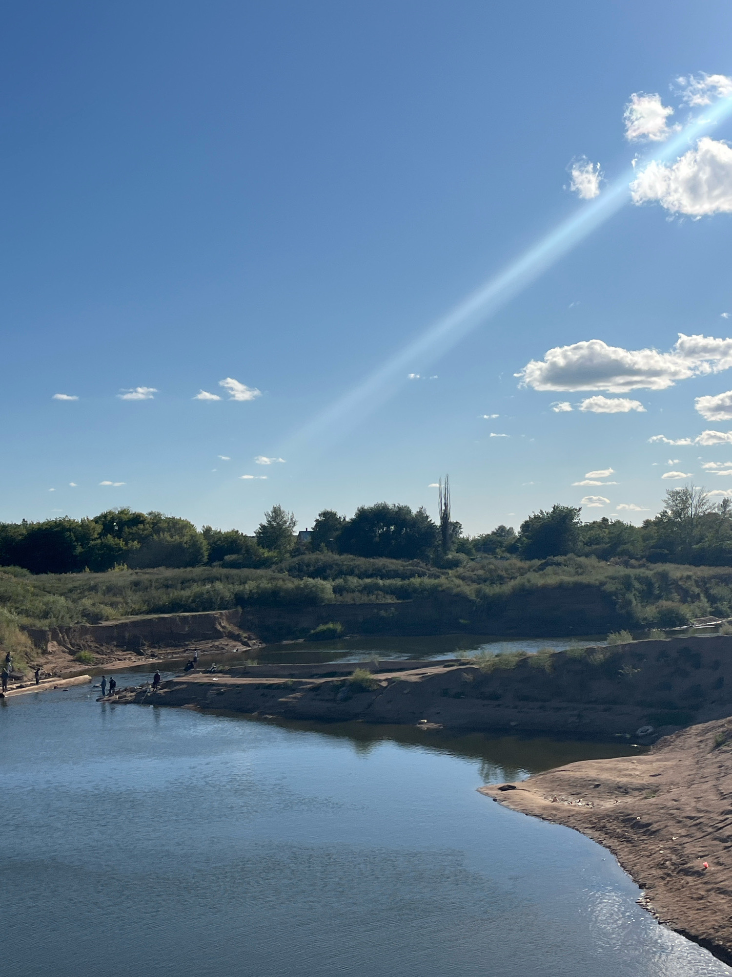 The image shows a wide river with sandy banks under a bright blue sky dotted with white clouds. A prominent sunbeam streaks across the sky from the upper right. On the left bank of the river, several people are visible, some standing and appearing to be fishing. The background consists of dense green foliage and trees, with a few taller, thinner trees standing out. The river appears calm, with reflections of the sky and surrounding vegetation on its surface. The scene is natural and appears to be a recreational area. This image does not provide enough information to determine a specific location in the city of Astana, Kazakhstan.