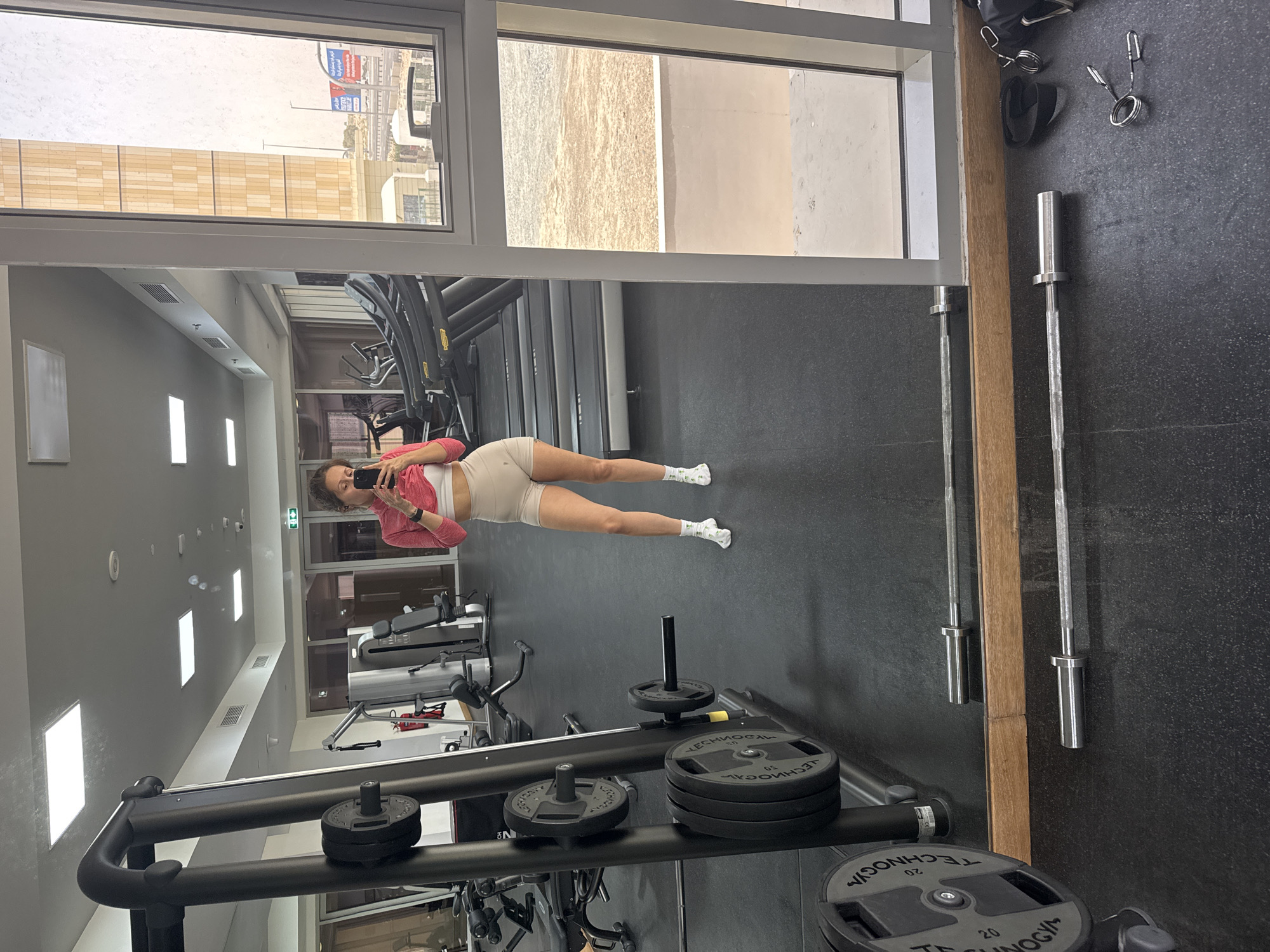 A mirrored reflection of a woman in a gym setting is captured within the frame. The gym appears to be well-lit with various fitness machines and equipment visible. The woman is standing facing the mirror and using her phone to take a photograph. She is dressed in athletic attire. Outside the gym windows, an urban landscape is seen, suggesting a city location. One of the windows reflects the Dubai skyline. Exercise equipment is present, with weights stacked on a machine and barbells hanging on the wall.