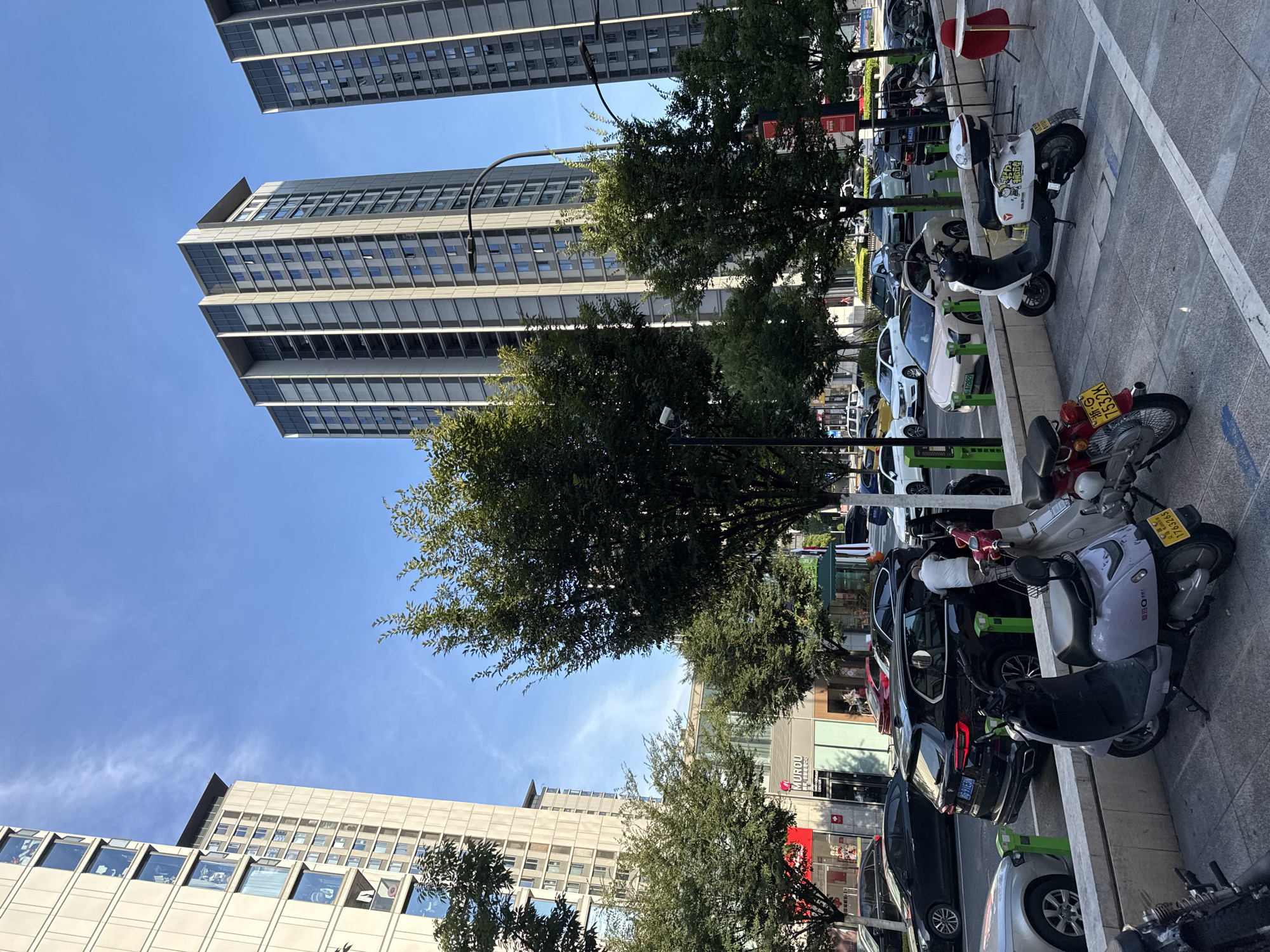 In the city of Yiwu, China, a street scene unfolds under a clear blue sky with wispy clouds. Tall, modern apartment buildings with numerous windows line the background, interspersed with mature trees. In the foreground, a street is lined with parked cars and several scooters. A person in a white shirt is seen interacting with the open trunk of a black SUV. The pavement is paved with square tiles, and a low concrete barrier separates the street from the sidewalk. A few businesses with storefronts are visible behind the trees. The overall impression is one of urban activity and daily life.
