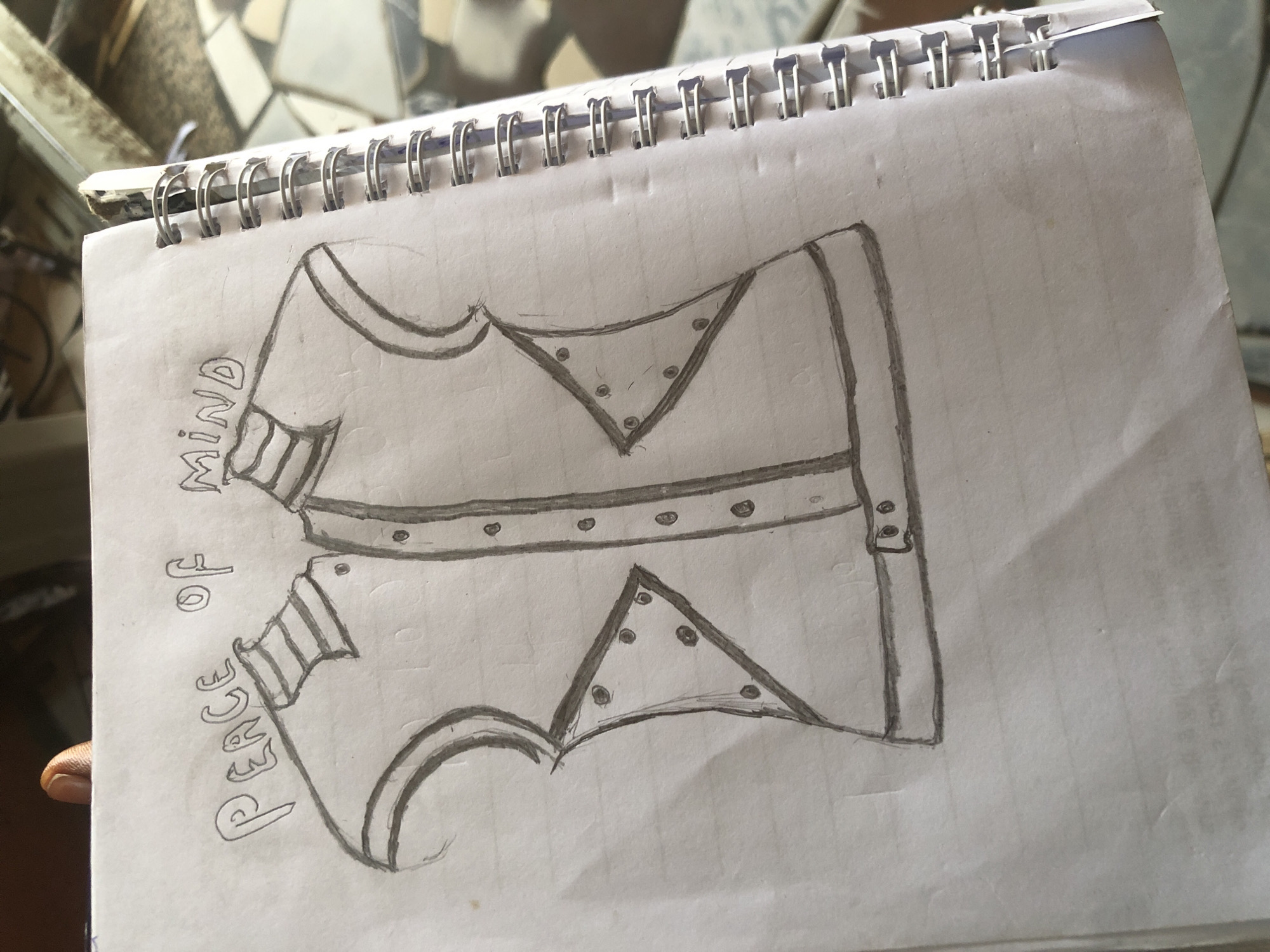 The image shows a hand-drawn sketch of a vest on a spiral-bound notebook. The vest is drawn in a simple, outline style with button details down the front. Above the vest, written in block letters, are the words "PEACE OF MIND". The notebook is open to a page with faint horizontal lines. A person's finger is visible at the top left of the image, holding the notebook. The background is blurred and appears to be an outdoor setting with tiled surfaces, suggesting it could be a patio or walkway in Ado, Nigeria.