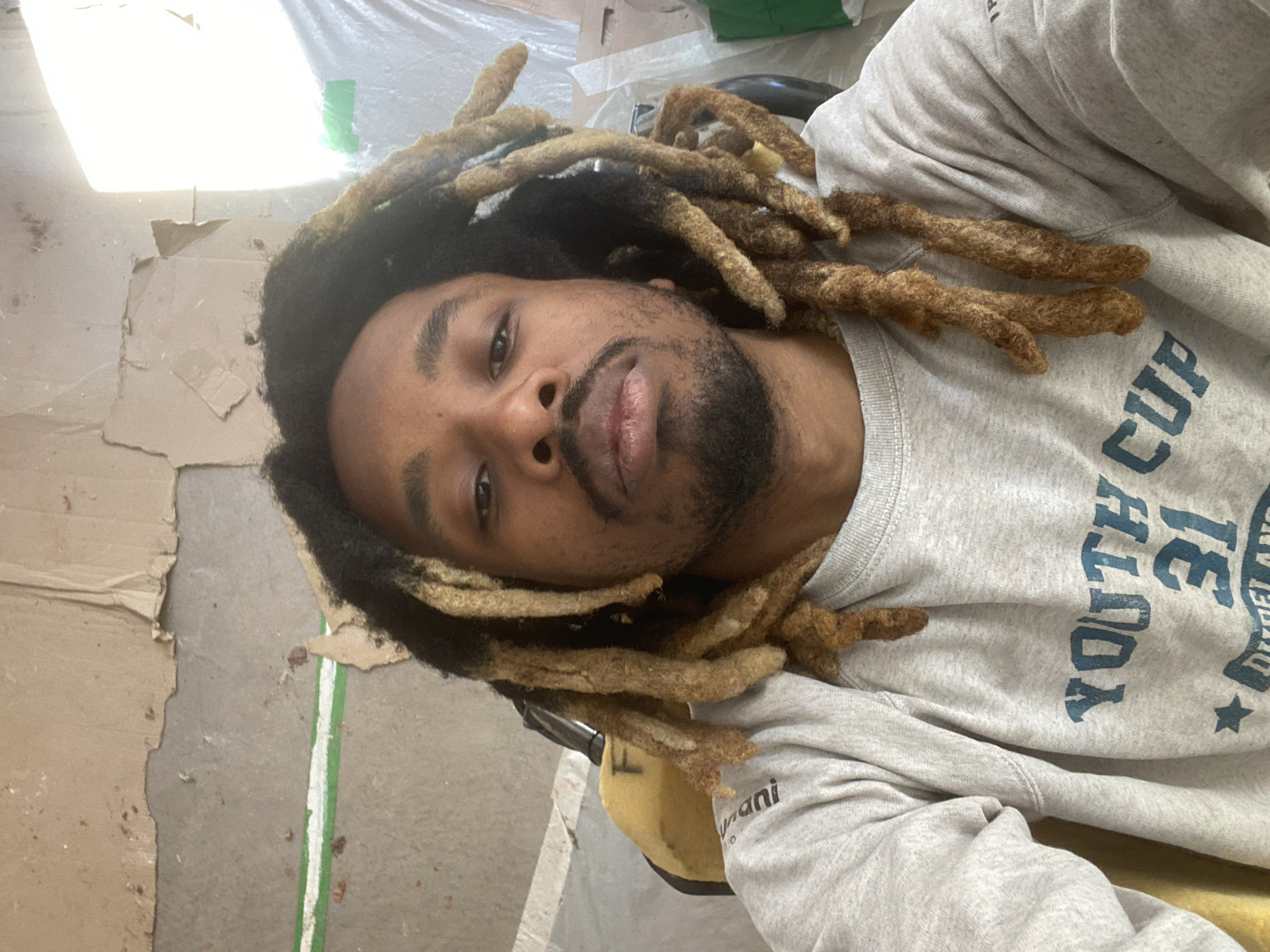 The media file is a vertical, eye-level selfie depicting a young man with dark skin, a beard, and distinctive dreadlocks. His dreadlocks are dark at the roots and gradually transition to a lighter, caramel-blonde color towards the ends. He looks directly into the camera with a neutral expression.
The man is wearing a light grey crewneck sweatshirt. Prominently displayed on the front of the sweatshirt in blue, distressed-style lettering are the words "YOUTH CUP" followed by the number "31." Partially visible text below includes what appears to be "DIESEL" or similar branding, and the suffix "mani" (likely from a word like "Humanity" or a brand name) is seen on his left arm area.
The background suggests an indoor, informal setting, possibly a workspace, storage area, or a room undergoing renovation. Surfaces are covered with light brown cardboard sheets, some appearing torn or aged. A bright, intense light source, likely an overhead lamp or window, is visible in the upper left corner, casting a strong glare. White plastic sheeting is also present in the background. The floor beneath appears to be a light grey concrete or industrial surface, marked with a thin green and white stripe. The overall atmosphere is utilitarian and somewhat unkempt. Although not explicitly visible in the image, this photo was taken in Paris, France.