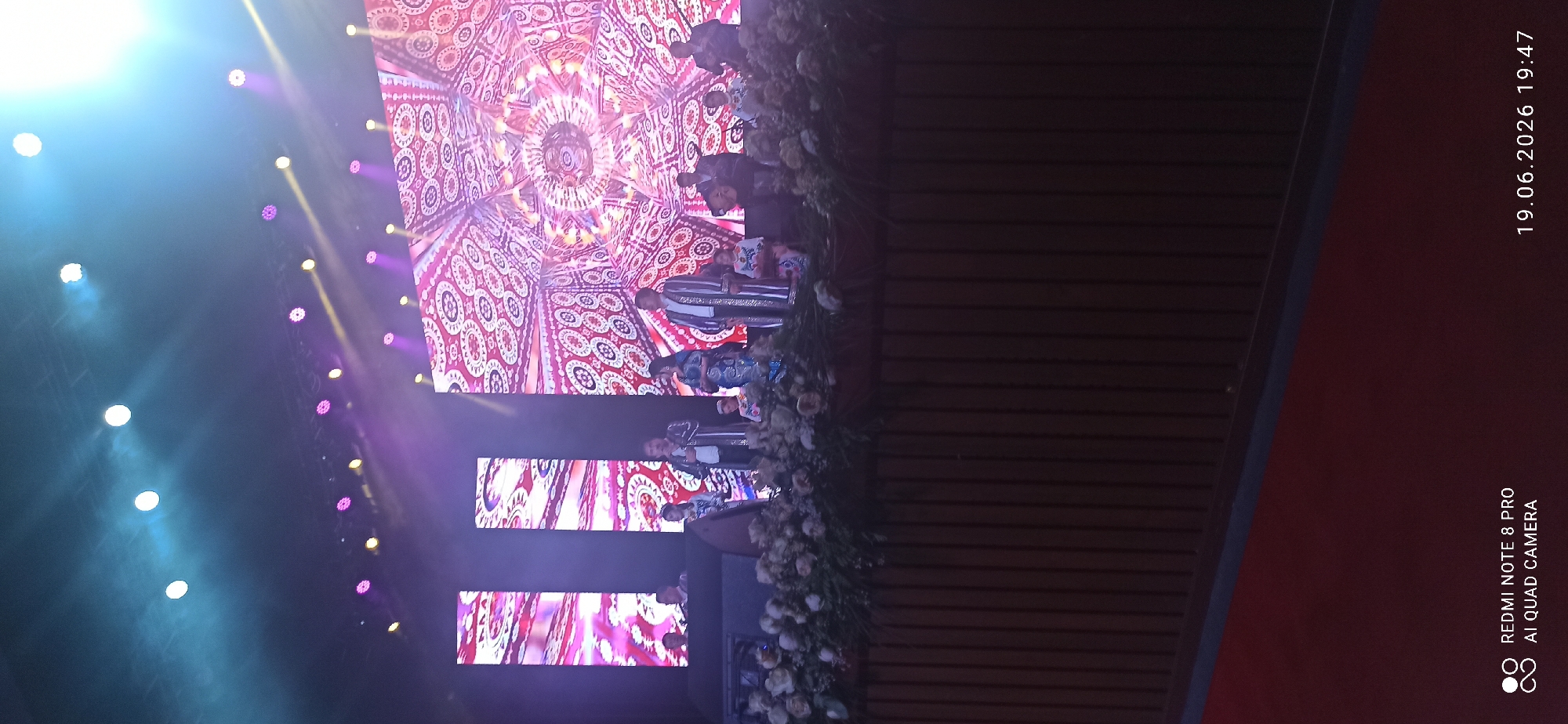 A group of people are performing on a stage in front of a large LED screen. The screen displays a colorful, intricate pattern. Several individuals are dressed in traditional clothing and appear to be singing or speaking. One person is playing a guitar. The stage is decorated with flowers. The lighting on stage is bright and dynamic, with spotlights and colored lights. The image was taken in Unknown, Unknown at 19:47 on June 19, 2026. The bottom left corner of the image contains text that reads "REDMI NOTE 8 PRO AI QUAD CAMERA".