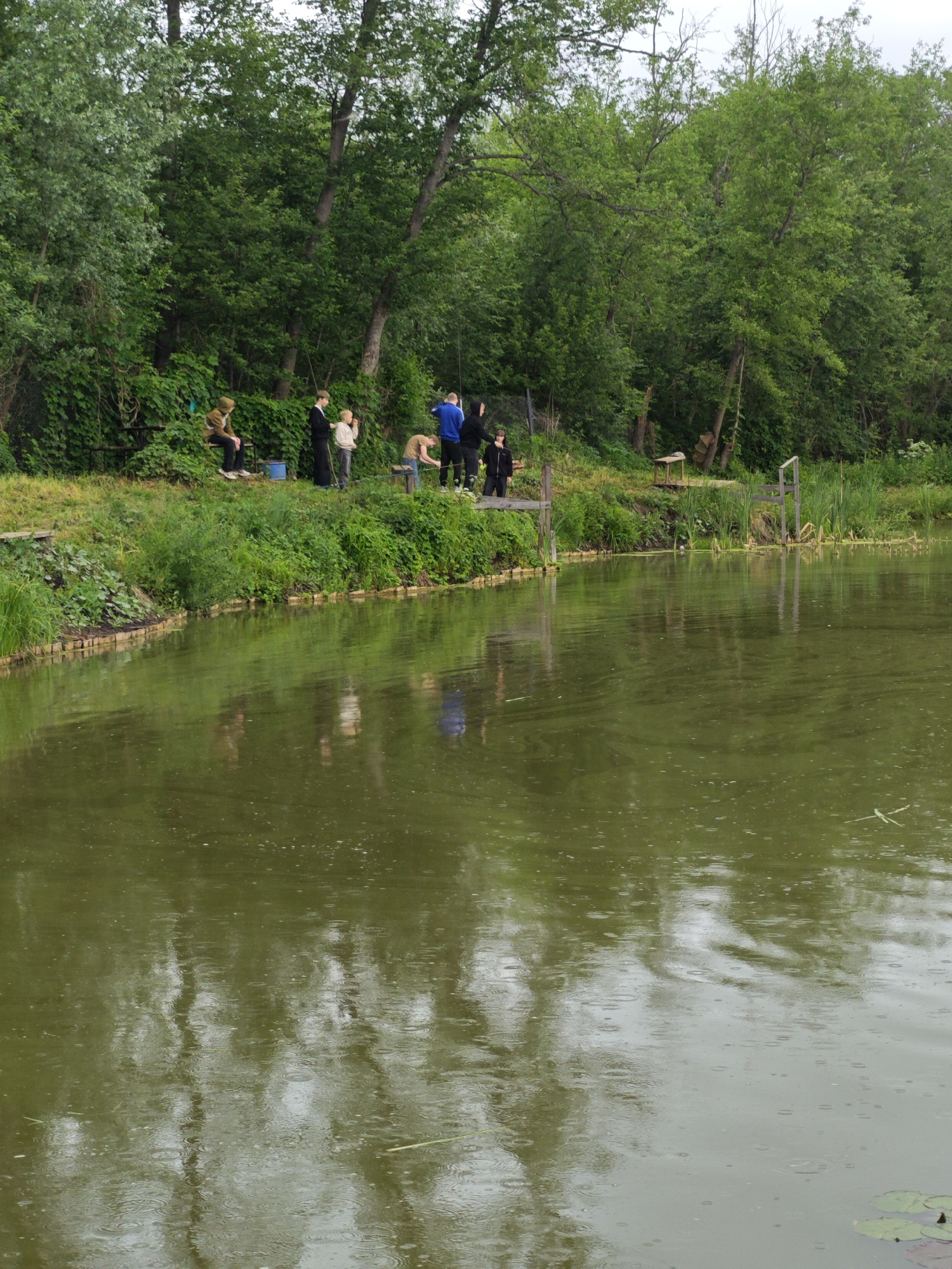The image captures an outdoor scene at the edge of a body of water in Malyi Bukor, Russia. The water, likely a pond or calm river, is a murky greenish-brown and exhibits numerous small ripples and concentric circles on its surface, indicative of light rain. Reflections of the overhead trees and sky are visible on the water.
Along the immediate bank of the water, a low retaining wall constructed from horizontal wooden planks is present. The bank itself is covered in lush green grass and dense vegetation, including numerous shrubs and a thick line of deciduous trees that form a verdant background. A simple wire fence is visible on the middle-left section of the bank.
Approximately eight individuals are positioned along the bank. From left to right:
1. An individual, mostly obscured by the left frame, appears to be wearing dark clothing.
2. A person in a brown or khaki hooded jacket and dark pants is seated on a low wooden bench. A blue bucket rests on the ground near their feet.
3. A person in a long, dark coat or robe stands facing right, looking towards the water.
4. A younger individual, possibly a child or teenager, wearing a light-colored top and dark pants, stands looking towards the water.
5. A person in a light tan top and dark pants is leaning forward over the water's edge.
6. An individual in a blue long-sleeved shirt and dark pants stands upright, looking towards the water.
7. A person in a dark hooded jacket and dark pants stands to the right of the individual in blue.
8. Another individual, partially obscured on the far right, is wearing dark clothing.
Towards the right side of the frame, a rustic wooden structure, possibly a small dock or platform, extends slightly over the water, supported by wooden posts. A few green lily pads are visible on the water surface in the bottom right corner. The overall atmosphere suggests a damp, overcast day in a natural, forested environment.