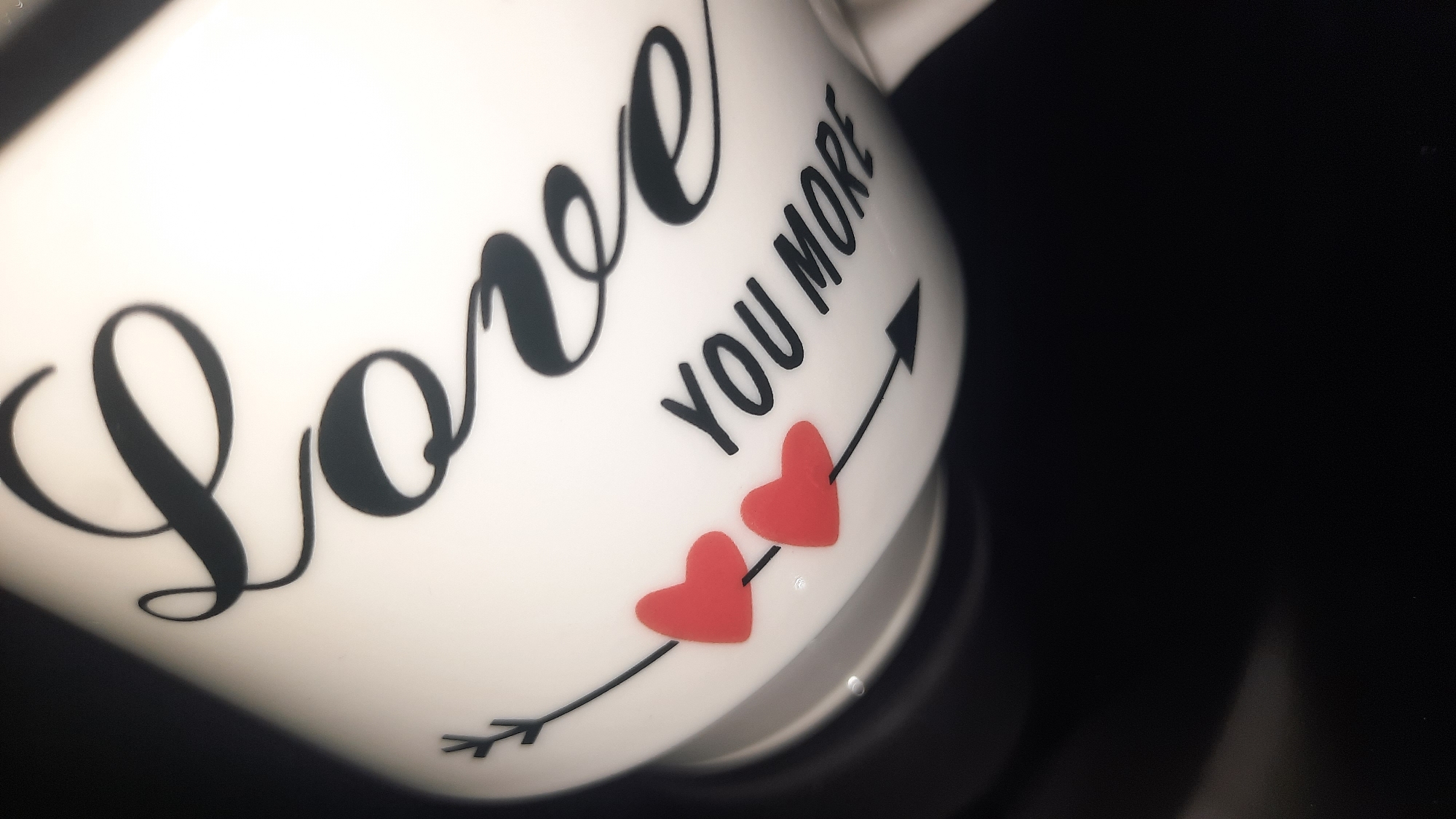 The image shows a close-up of a white ceramic mug with black lettering that reads "Love you more". Two red heart decals are placed near the word "Love". An arrow is depicted as part of the design, pointing from left to right, passing through the hearts. The background is dark and out of focus. There is no discernible location in the city of Шклоў, Belarus.