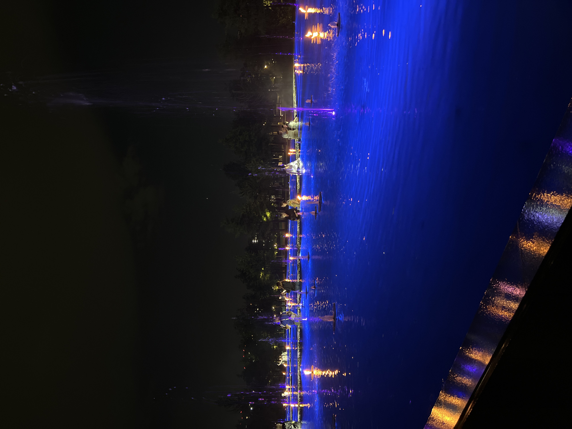 The image shows a fountain at night in the city of Krasnodar, Russia. The fountain is illuminated with blue lights, and there are people standing in and around the fountain. Some of the people are holding candles, which are also lit and reflected in the water. The fountain is surrounded by trees, and in the background, there are some buildings and streetlights. The scene is dimly lit, with the fountain being the main source of light. The water in the fountain appears to be calm, with ripples on the surface.