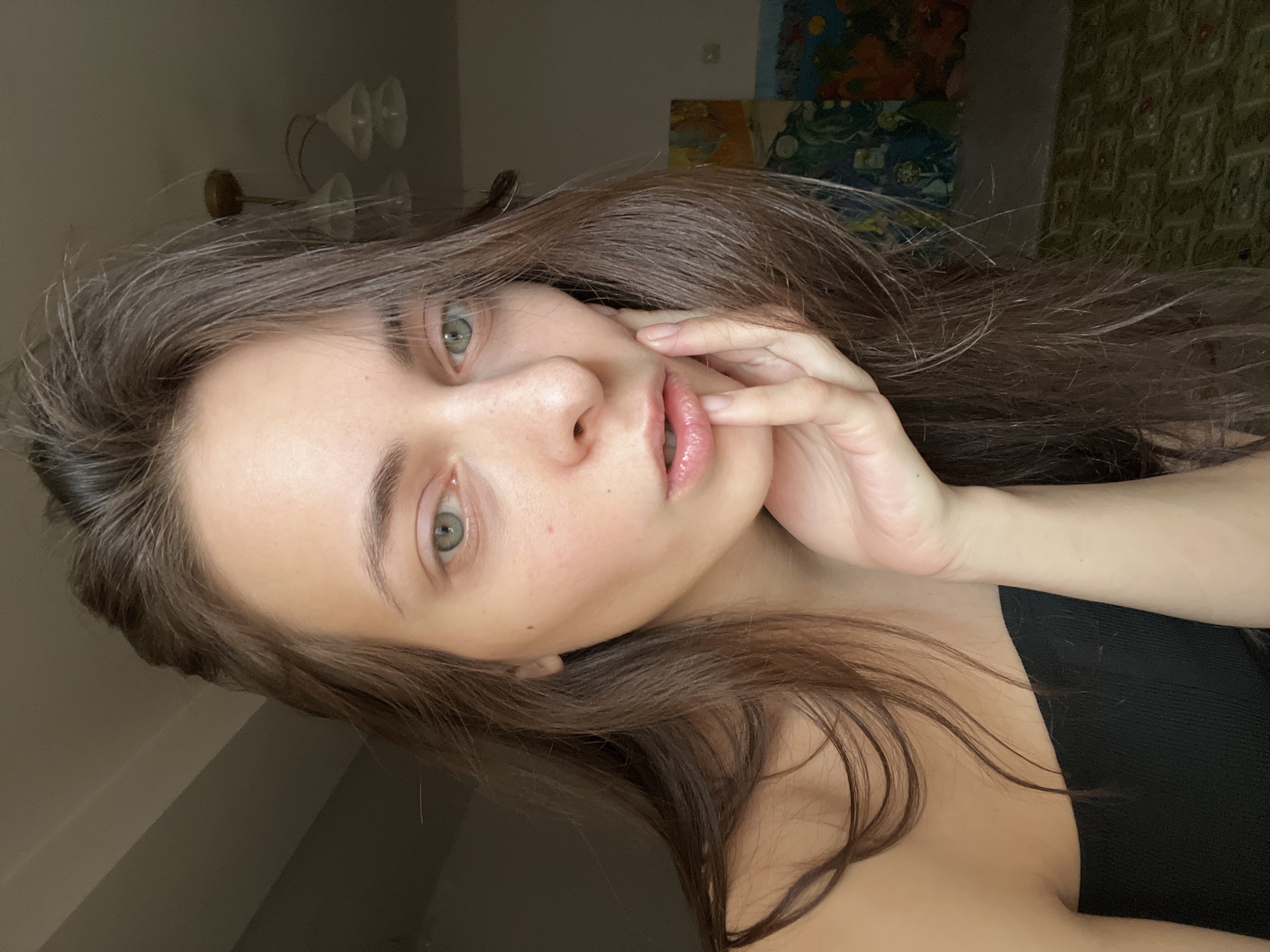 A young woman with long brown hair is seen in a close-up, slightly tilted shot. Her face is pale and features a light complexion, with light-colored eyes and glossy lips. Her hand is gently placed near her face. She is wearing a black top. The background is blurry, but what appears to be a lamp and some paintings can be seen. There is no verifiable information to place the subject at a specific location, such as Krasnoyarsk, Russia.