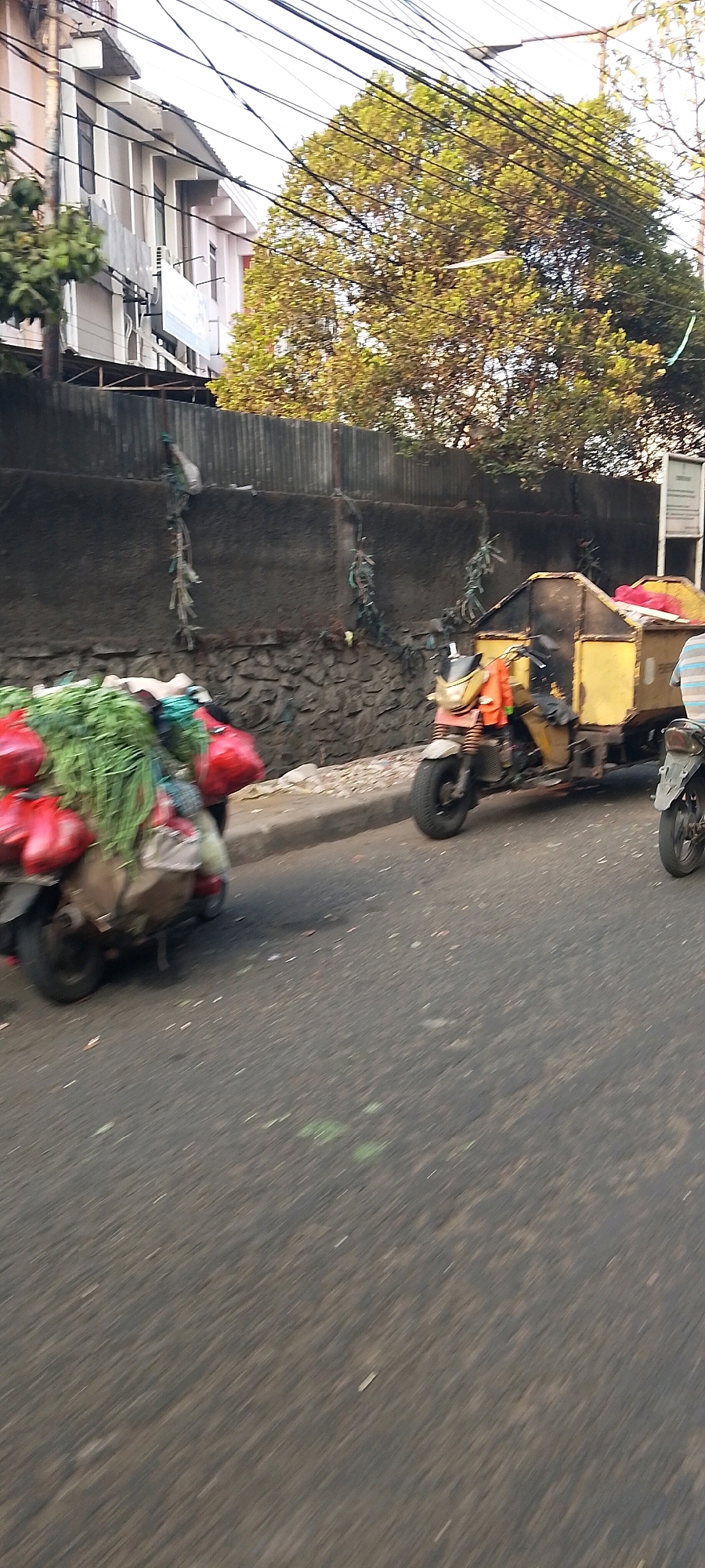 In Bekasi, Indonesia, a three-wheeled yellow motorized vehicle, possibly a waste or goods transport, is observed on the right side of the image. It is laden with a large, dark yellow container at the rear, suggesting it is a cargo vehicle. The vehicle is parked or moving slowly on a paved road, with its front wheel visible. To the left, another three-wheeled vehicle, possibly a motorcycle with a cargo attachment, is partially visible. This vehicle is loaded with a substantial amount of green vegetables and several red bags, suggesting it is used for transporting produce. The scene unfolds against a backdrop of a textured stone and corrugated metal wall, with trees and power lines overhead. A building with multiple windows and air conditioning units is situated behind the wall, indicating an urban setting. The overall image depicts a moment of daily activity on a street in Bekasi, likely related to commerce or transportation of goods.