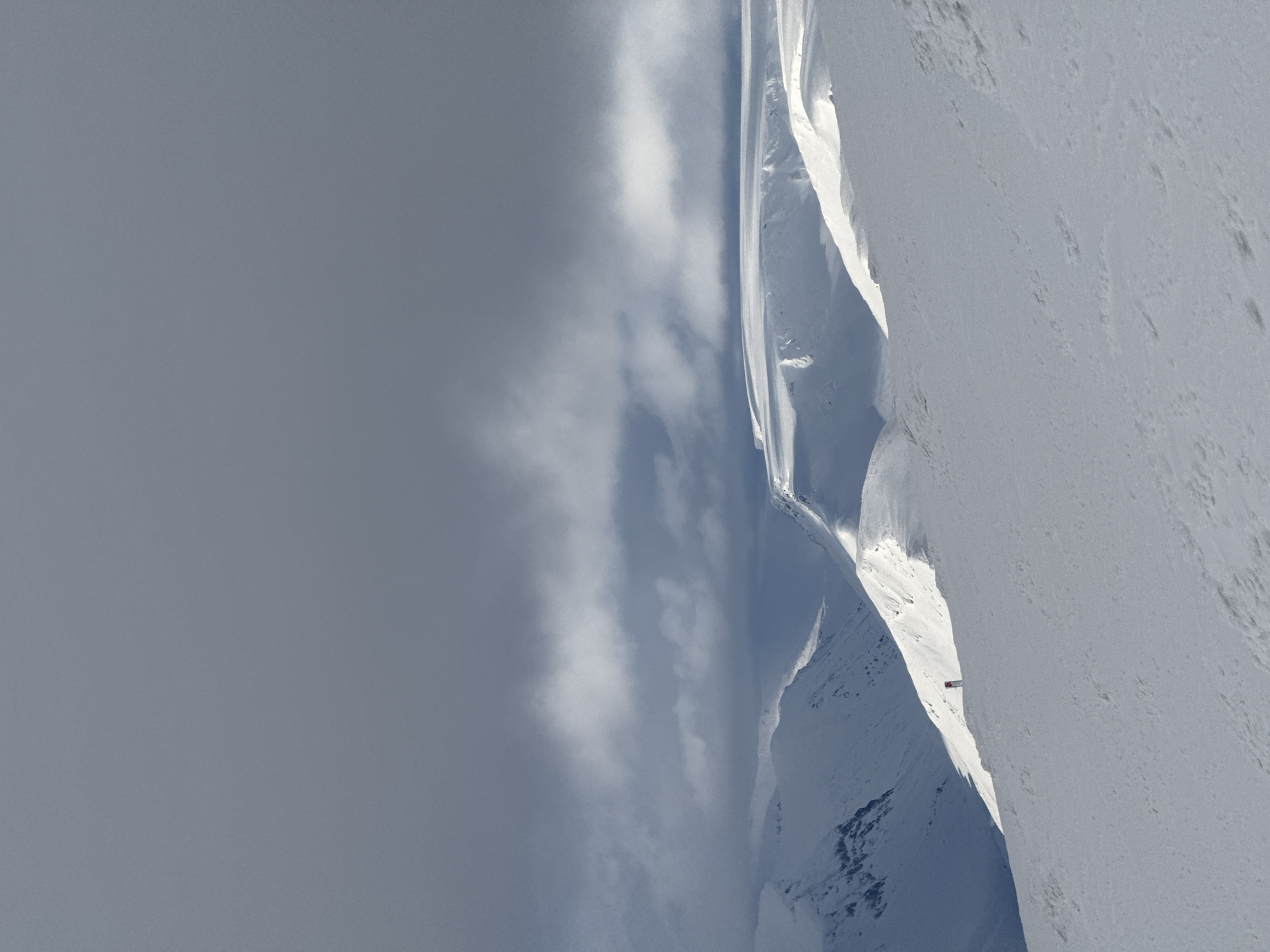 The image shows a snowy mountain range under a cloudy sky. The landscape is dominated by snow-covered peaks and ridges. A small, red structure is visible on one of the slopes. The image is rotated. The exact location is unknown.