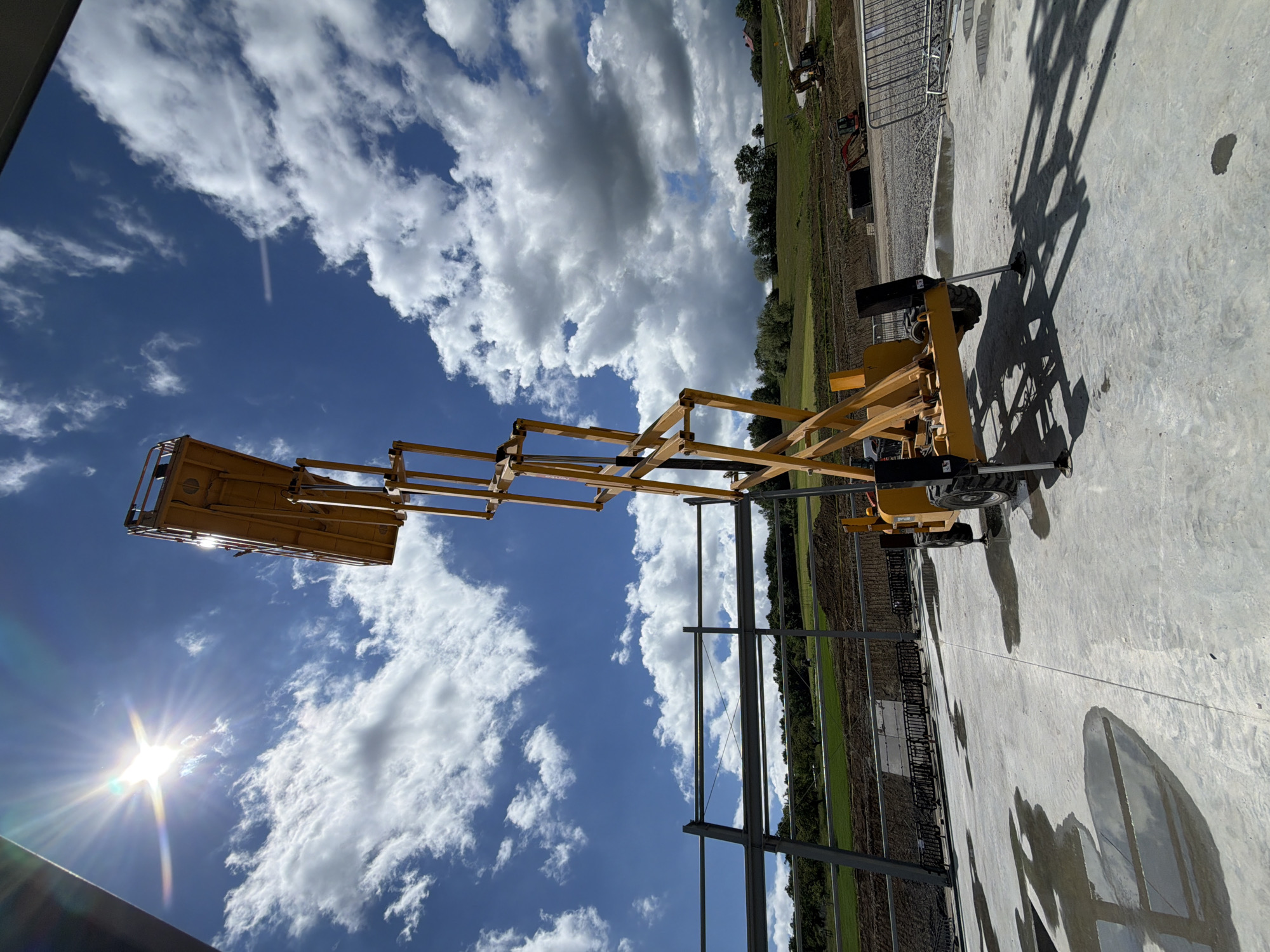 An extended yellow scissor lift is positioned on a concrete surface under a bright, sunny sky with scattered clouds. The scissor lift is partially in shadow, and its platform is extended upwards. In the background, a partially constructed building with metal beams is visible, along with a green, hilly landscape and a road. Several small excavators are present on the construction site. The scene appears to be a construction area in Janowice, Poland.