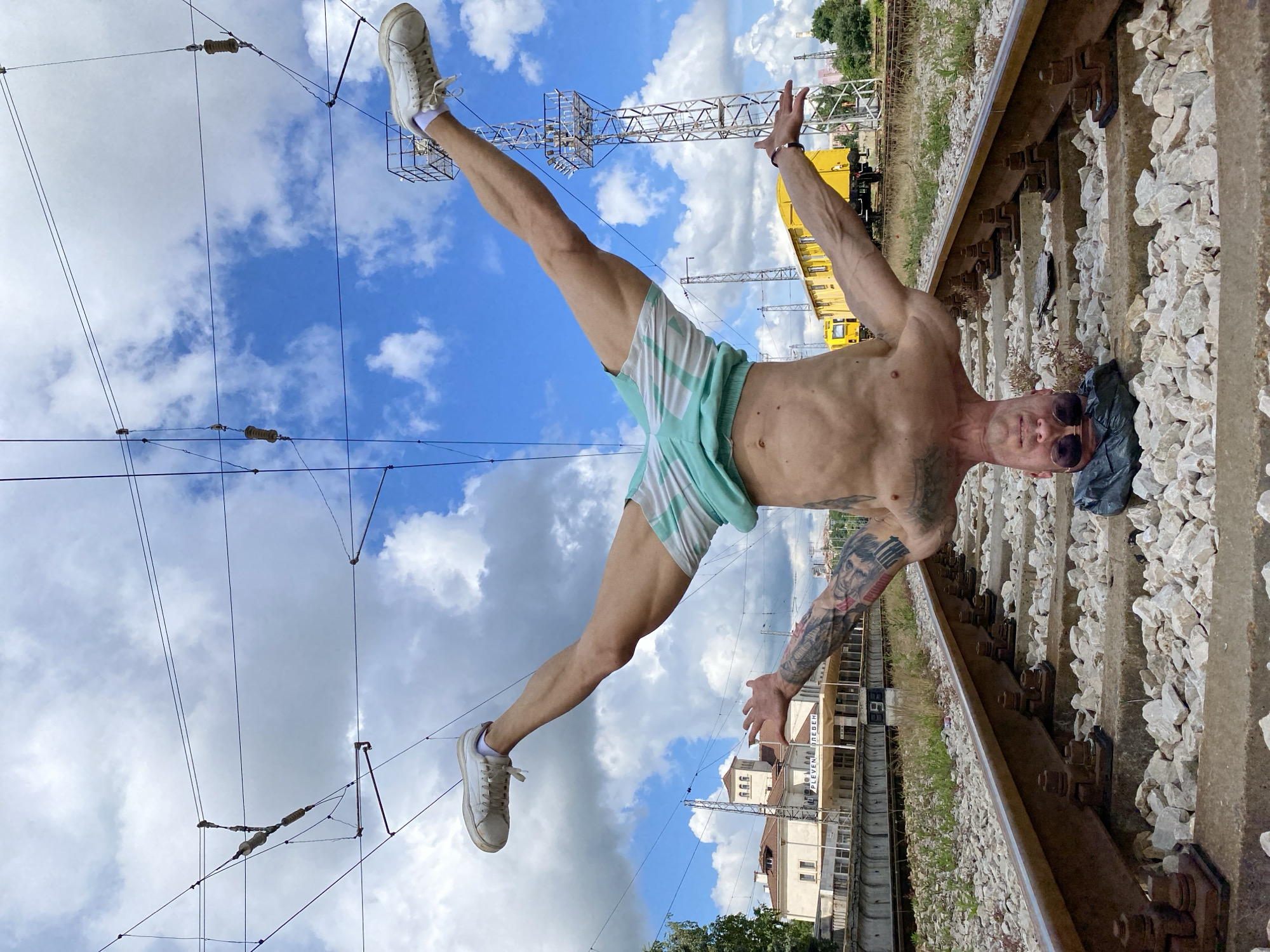 A shirtless man is performing a handstand on a set of train tracks in Pleven, Bulgaria. He is wearing turquoise and white striped shorts and white sneakers. His legs are spread wide apart, and his arms are extended outwards for balance. His head is resting on a dark bag on the ground, and he is wearing sunglasses. Tattoos are visible on his arms and torso. In the background, there is a train station building with Cyrillic signage that reads "PLEVEN". Yellow train cars and overhead electrical wires for the train are also visible. The tracks are surrounded by ballast and some dry grass. The sky is blue with scattered white clouds.