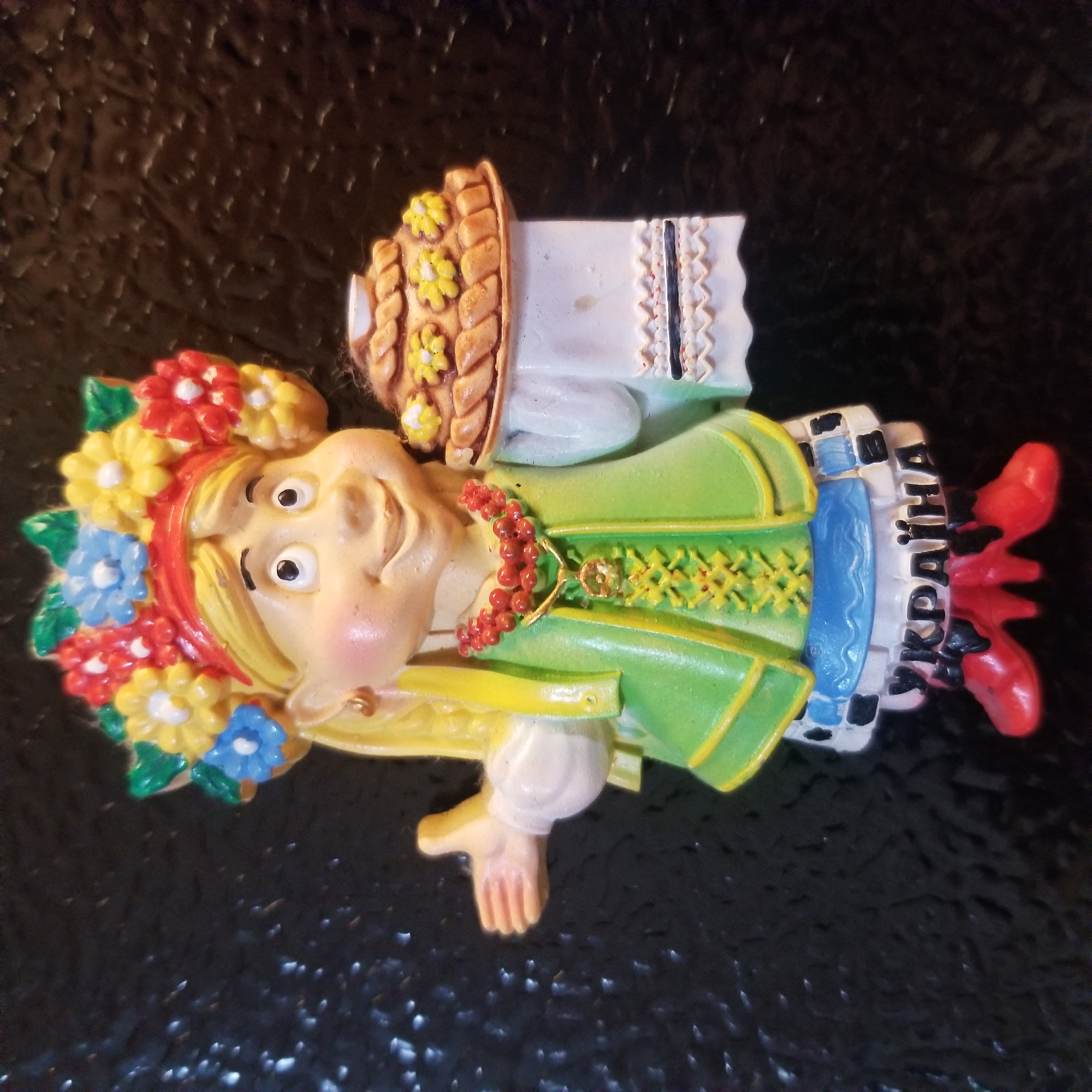 This is a photograph of a ceramic magnet shaped like a Ukrainian girl. The girl is wearing a traditional Ukrainian headdress adorned with colorful flowers and a red ribbon. She has blonde hair and a smiling face with rosy cheeks. She is dressed in a green vest with yellow trim and embroidered details, a blue skirt, and a white apron with a checkered pattern. She is holding a white cloth in one hand and a traditional Ukrainian pie, called a karavai, decorated with yellow flowers, in the other. The word "Україна" (Ukraine) is written in black letters on her apron. She is wearing red shoes with black bows. The magnet is set against a dark, textured background that appears to be a refrigerator or similar magnetic surface. The location is not identifiable from the image itself, and therefore cannot be specified as Virginia Beach, United States of America.