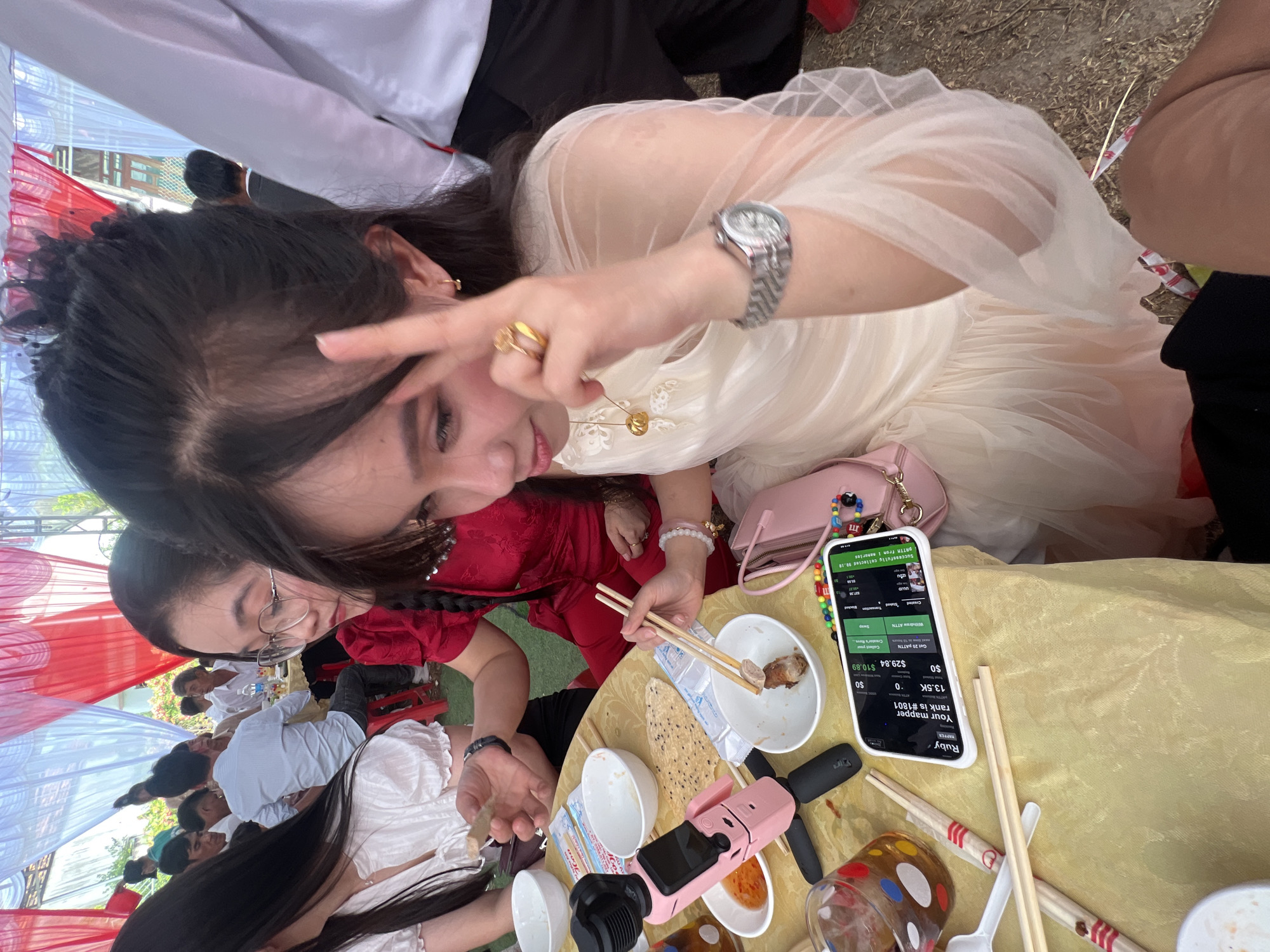 The image depicts a lively social gathering taking place outdoors in Tay Son Commune, Vietnam, beneath a festive canopy of red and white fabric. The central figure is a young woman with long, dark hair, wearing an elegant light-colored, possibly cream or white, flowing dress. She is smiling and making a peace sign with her right hand, displaying a silver watch, gold rings, and a delicate gold necklace, conveying a cheerful and relaxed mood.
In front of her, on a table covered with a yellow cloth, a white smartphone is prominently displayed. Its screen shows an application with various details: "Ruby," "RAPPER," "Your mapper rank is #1801," "13.5K," "0," "$0," "$0," "$29.84," "$10.89," "Get 20 PATTN," "Collect your Creator's Reve," "Withdraw ATTN," "Swap," and "Successfully collected 90.18 PRTTH from I nenories." Adjacent to the phone are several white bowls, some containing food remnants, along with wooden chopsticks, a pink selfie stick or tripod, and a clear glass decorated with colorful polka dots. A small pink handbag also rests on the table.
To the left, another woman in a red outfit is partially visible, actively eating with chopsticks. In the blurred background, numerous other individuals, primarily men in light-colored shirts, are seated at additional tables, participating in the event. The scene suggests a casual daytime meal or celebration, characterized by communal dining and friendly interaction.