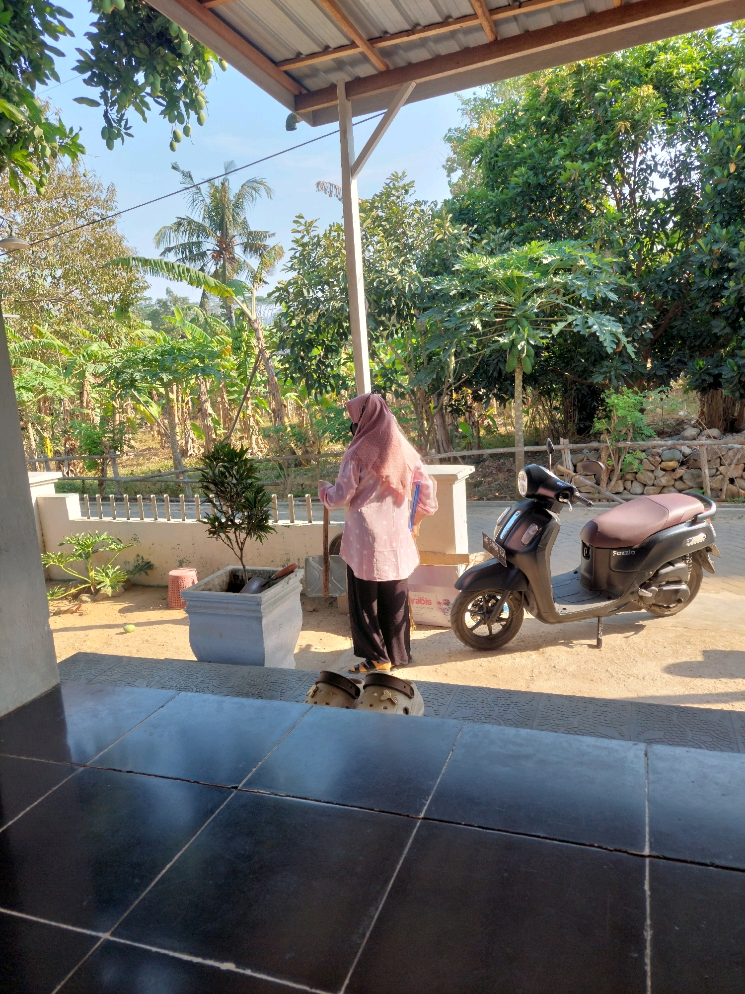 The image depicts an outdoor scene, likely a residential area, possibly in or near Menadi, Indonesia. A woman wearing a pink hijab and a pink long-sleeved top over black pants stands facing away from the camera. She is holding a blue folder. To her right, a black scooter with a brown seat is parked. The scooter has the brand name "FAZZIO" visible on its side and a license plate with text that is not clearly legible. In the foreground, to the left, is a grey planter containing a small tree and gardening tools. Next to the planter, a red stool is visible. Behind the planter, a patterned mat or flooring is laid out. Further in the background, there are trees, foliage, and a rustic stone wall. The ground appears to be sandy or dirt. The overall environment suggests a warm, sunny climate. The view is from under a covered patio or porch area, with a corrugated metal roof supported by a wooden beam visible at the top of the frame.