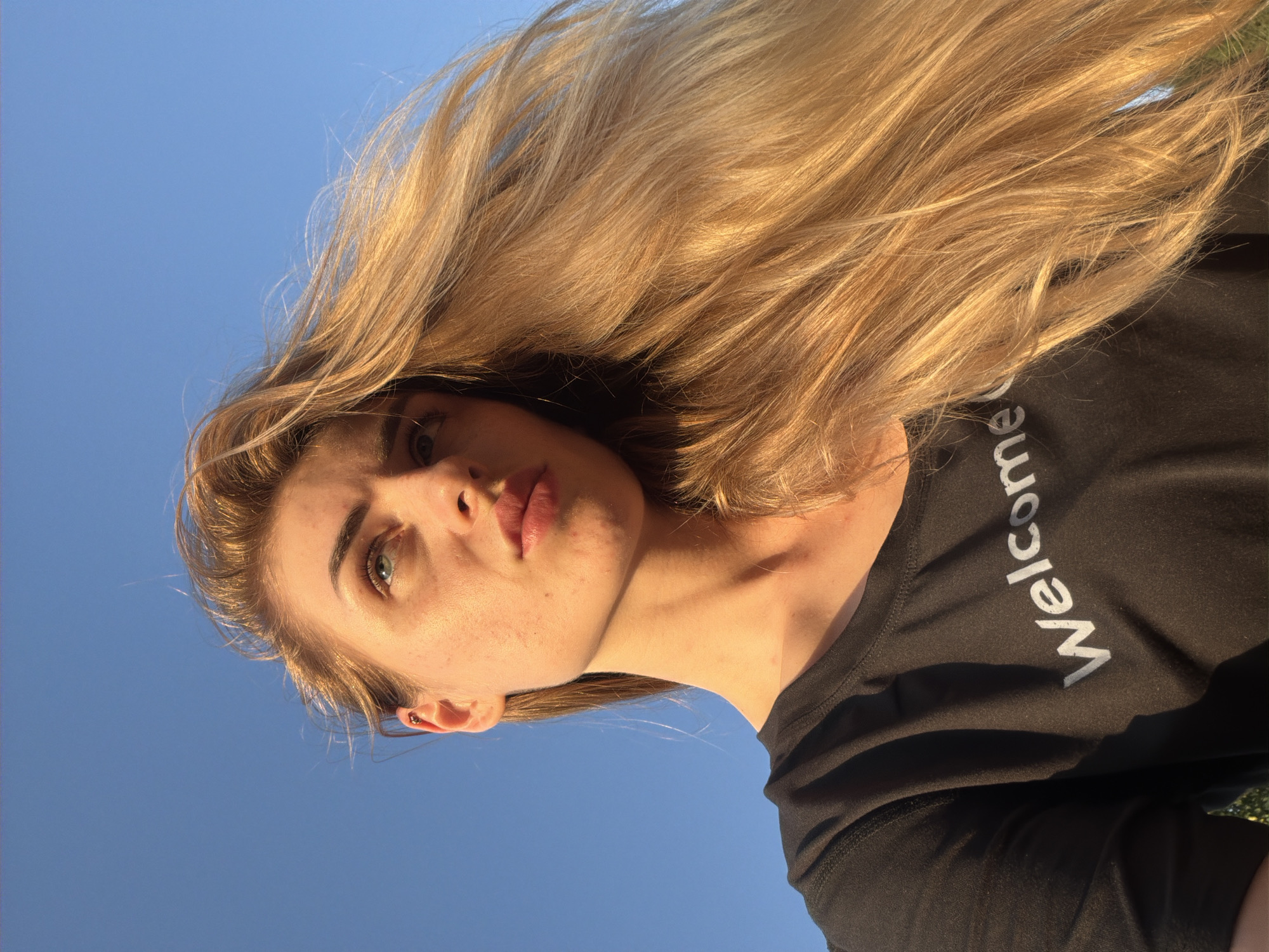 A medium shot shows a young Caucasian woman with blonde hair and blue eyes against a clear blue sky. She is looking upwards and to the left, with a slight furrow in her brow. Her blonde hair, which is long and windswept, frames her face and spills over her right shoulder. She is wearing a black t-shirt with the word "Welcome" printed in white letters across the chest. The sunlight illuminates the side of her face and hair, creating a warm glow. The lower part of the image shows a bit of her neck and shoulder, and the texture of the t-shirt fabric. The background is a uniform, cloudless blue sky. The image appears to have been taken outdoors, possibly during the golden hour due to the lighting.
