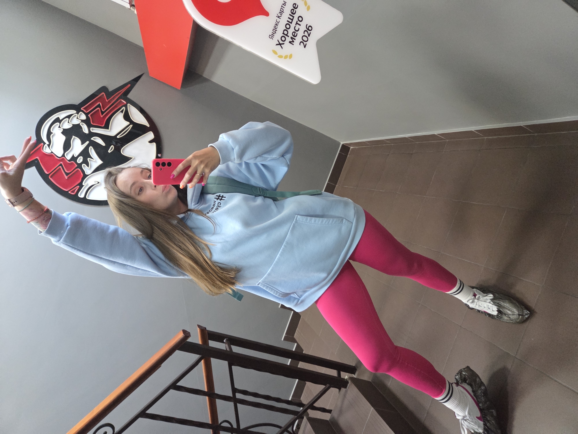A young woman with long, light brown hair is taking a selfie in a well-lit, indoor stairwell. She is wearing a light blue oversized hoodie with a kangaroo pocket and a teal backpack slung over her left shoulder. Her right arm is raised above her head, and her left hand is holding a pink smartphone, angled to capture her reflection. She is wearing bright pink leggings and white socks with black stripes, paired with white athletic shoes that have clear plastic covers over them. Her expression is a playful pout.
The stairwell features a dark wooden handrail and dark metal railings against a grey wall. The steps appear to be made of brown tile. In the background, a stylized logo with red, black, and white elements is visible on the grey wall, resembling a face with lightning bolts. To the right, a white sign with a red location pin graphic and text in Russian reads "Яндекс Карты Хорошее место 2026," which translates to "Yandex Maps Good Place 2026." The overall environment suggests a modern building, potentially a gym, a recreation center, or a public facility. The presence of the Yandex Maps sign indicates the location is likely in Russia, and specifically, given the context provided, Birobidzhan.